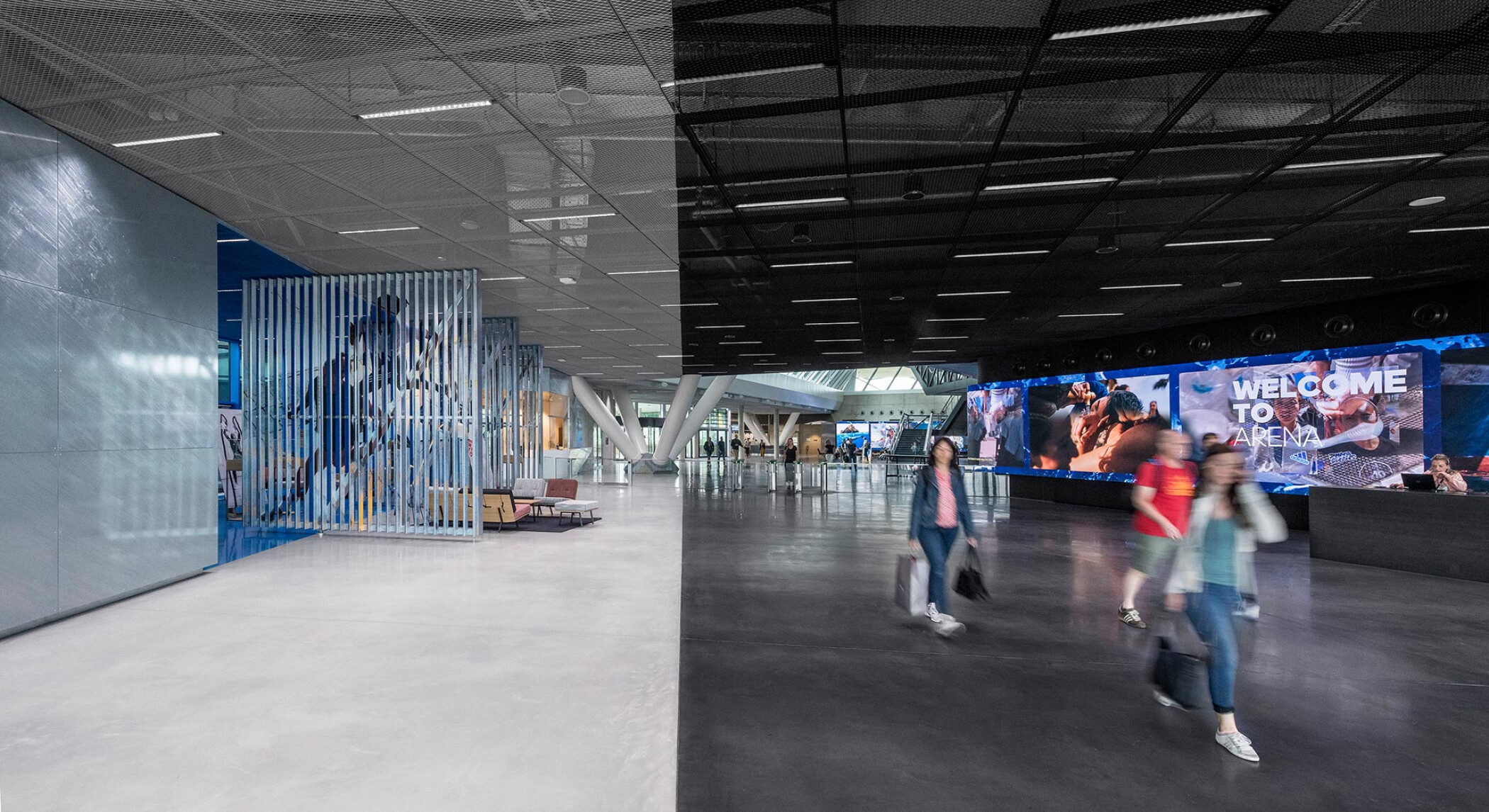 Project by Behnisch Architekturbuero, adidas ARENA. Large indoor lobby with open floor, people walking, reception desk, and a long digital screen displaying “Welcome to Arena.”