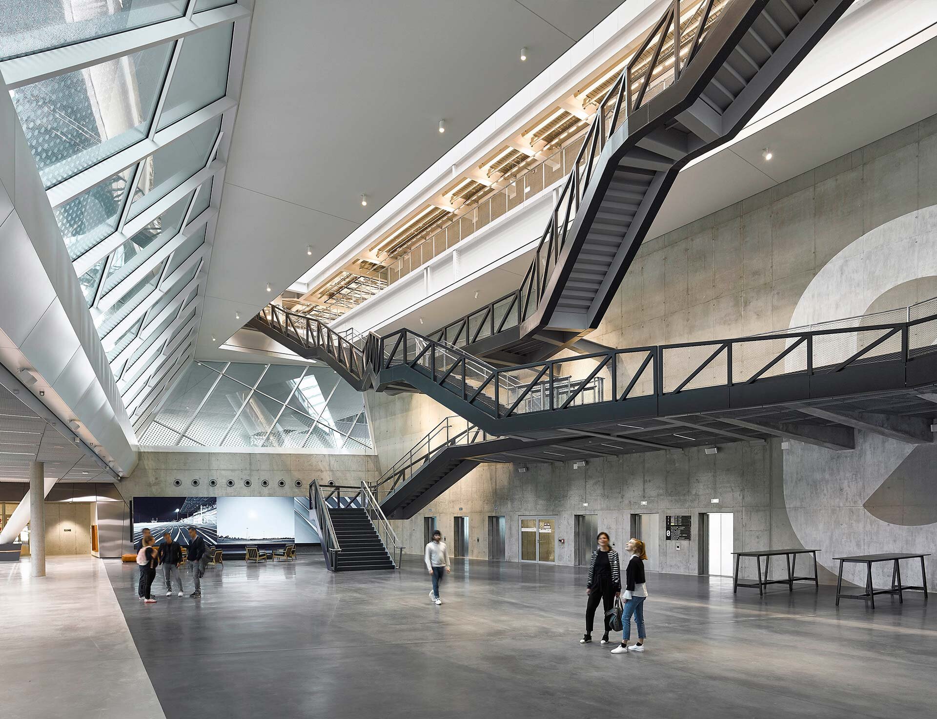 Project by Behnisch Architekturbuero, adidas ARENA. Large open atrium with exposed concrete walls, stairs and walkways, skylights and people in the entrance area.