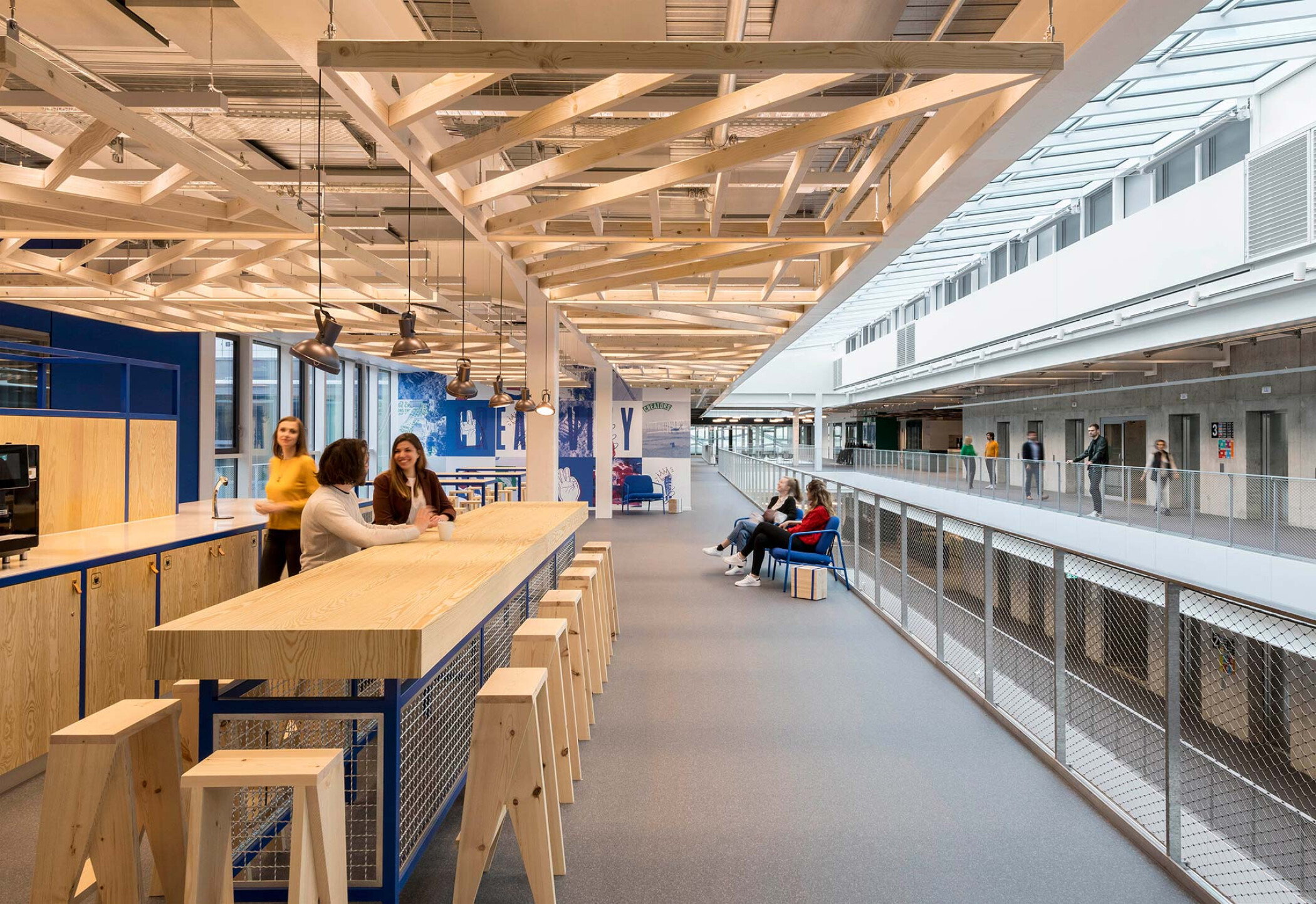Project by Behnisch Architekturbuero, adidas ARENA. Interior corridor with timber ceiling structure, long high table and stools. People are socializing in this area. On the right side there is a balcony walkway overlooking atrium.