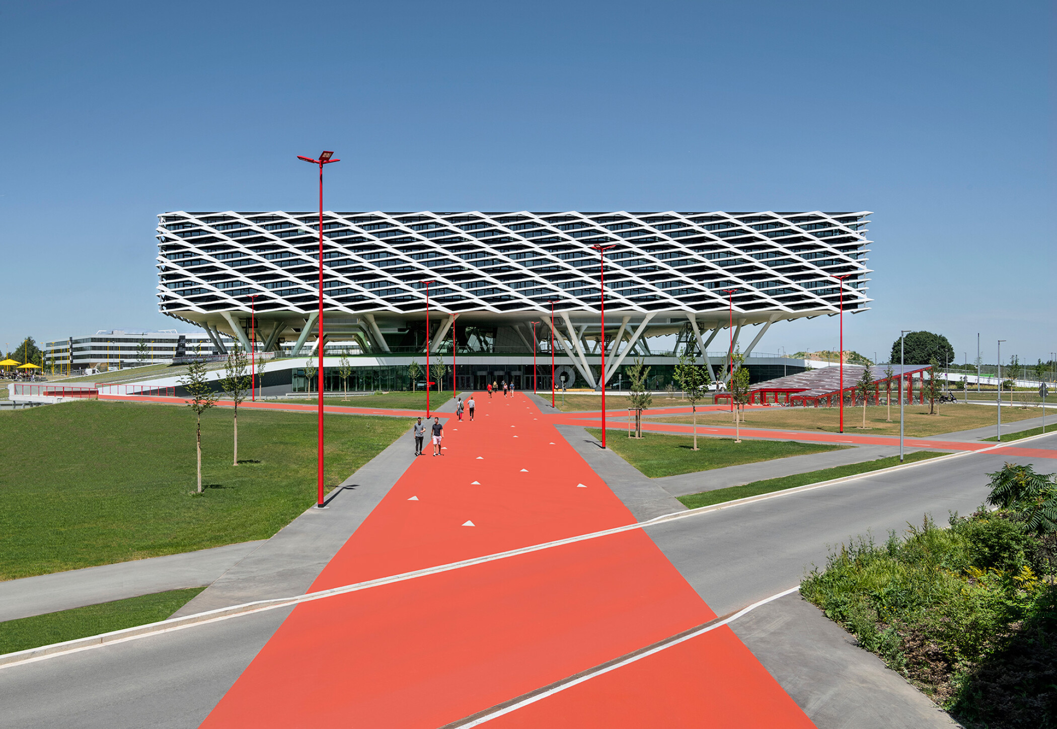Project by Behnisch Architekturbuero, adidas ARENA. A large, modern building with a sloping, grid-like facade features it is a specially designed sun protection and shading system. The main building is elevated and stands on stilts. People walk along a wide red path leading to the entrance.