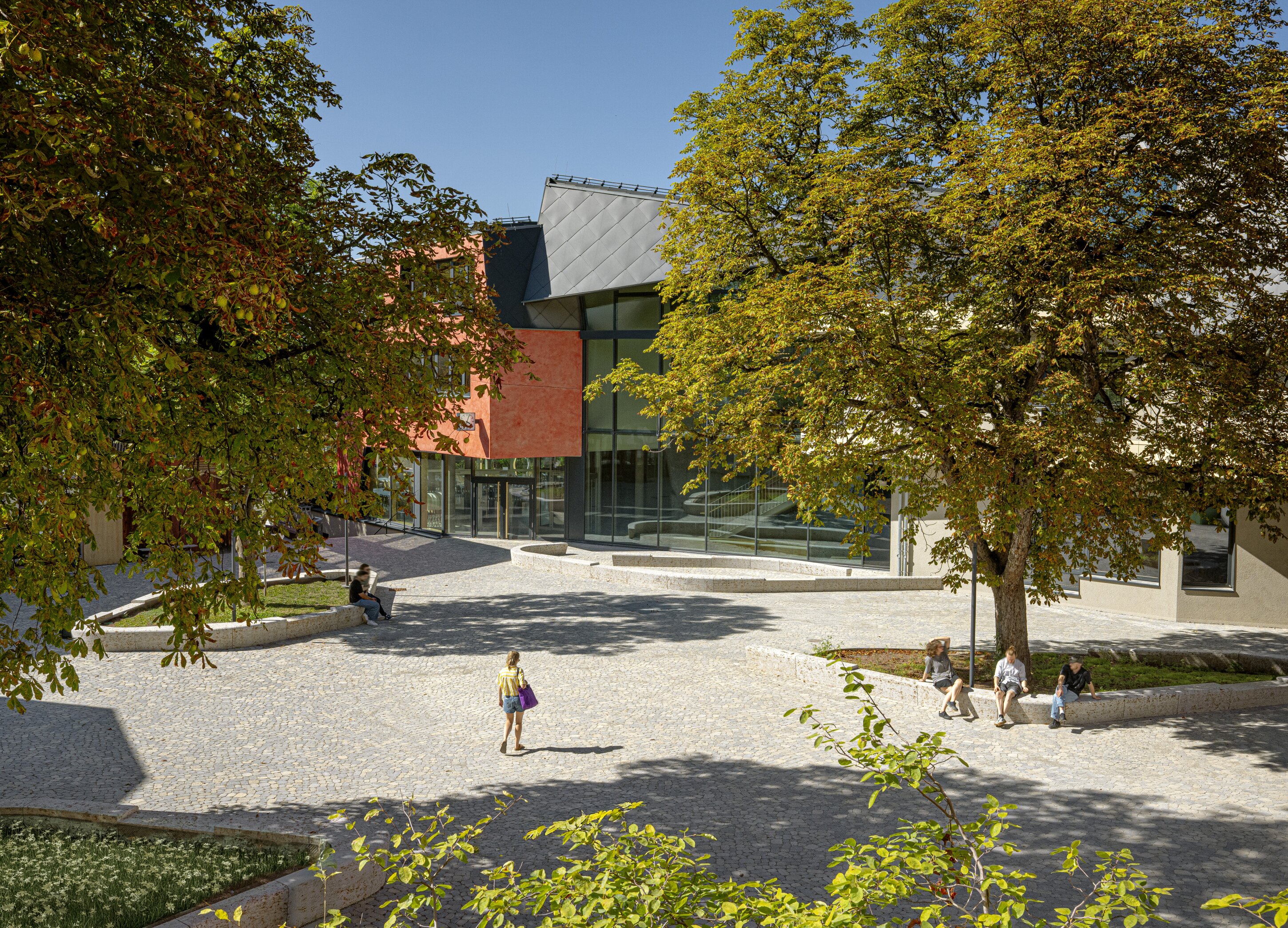 Project by Behnisch Architekturbuero, Freie Waldorfschule Uhlandshoehe - New School and Administration Building. Courtyard with trees and seating, people sitting and walking, in front of a modern building with glass facade and colorful exterior panels.