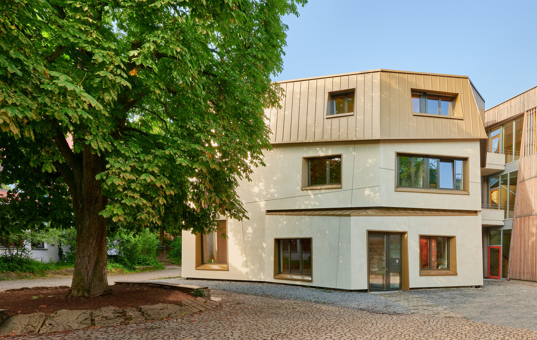 Project by Behnisch Architekturbuero, Freie Waldorfschule Uhlandshoehe - New School and Administration Building. Modern three-story building with light facade and metal roof, beside a large tree and paved courtyard.