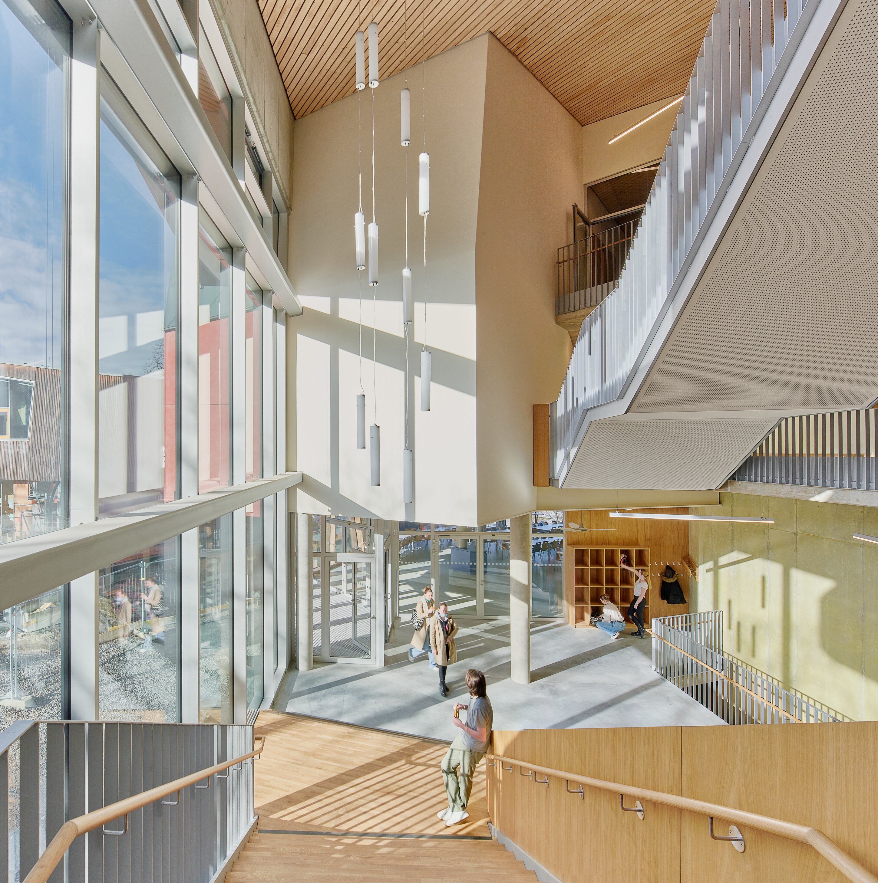 Project by Behnisch Architekturbuero, Freie Waldorfschule Uhlandshoehe - New School and Administration Building. Bright interior atrium with large windows, stairs, hanging lights, and people walking and sitting near an entrance and seating area.