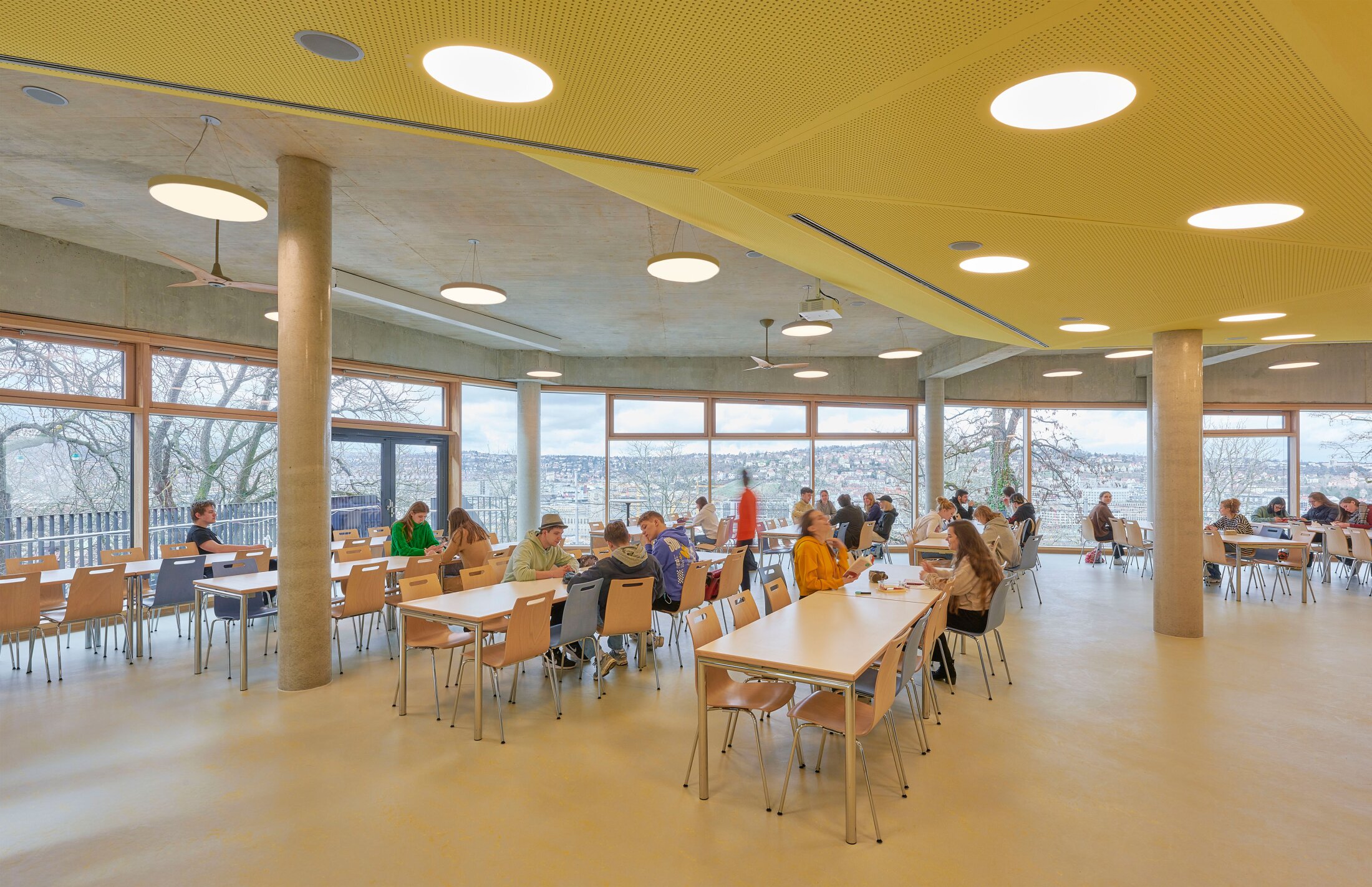 Project by Behnisch Architekturbuero, Freie Waldorfschule Uhlandshoehe - New School and Administration Building. Students seated at tables in a bright cafeteria with large windows, yellow ceiling panels, and city view outside.