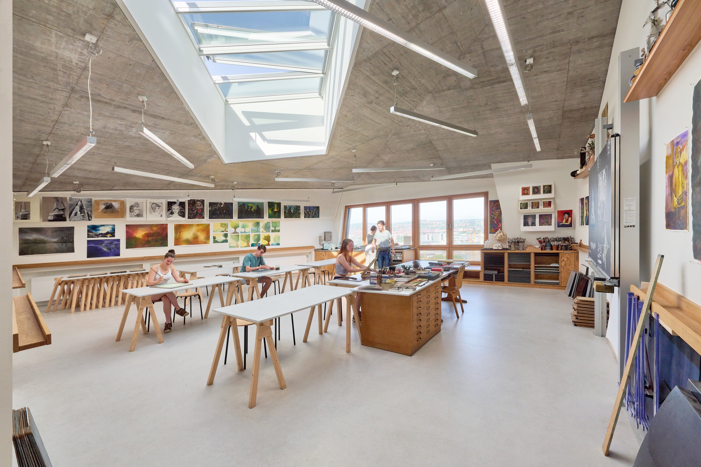 Project by Behnisch Architekturbuero, Freie Waldorfschule Uhlandshoehe - New School and Administration Building. Art classroom with students working at tables, artworks displayed on walls, large windows, and a skylight illuminating the space.