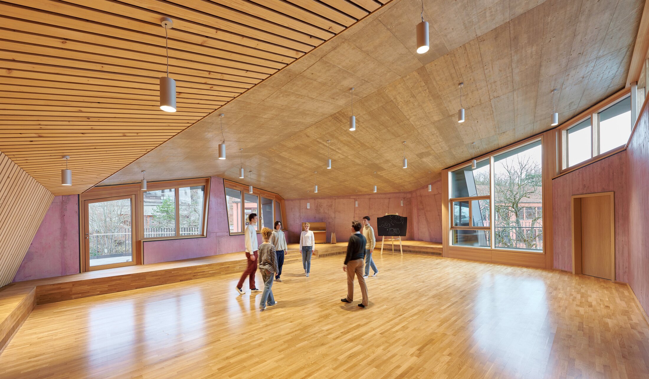 Project by Behnisch Architekturbuero, Freie Waldorfschule Uhlandshoehe - New School and Administration Building. Group of people standing in a large wood-floored room with sloped ceiling, windows, and a chalkboard in the background.