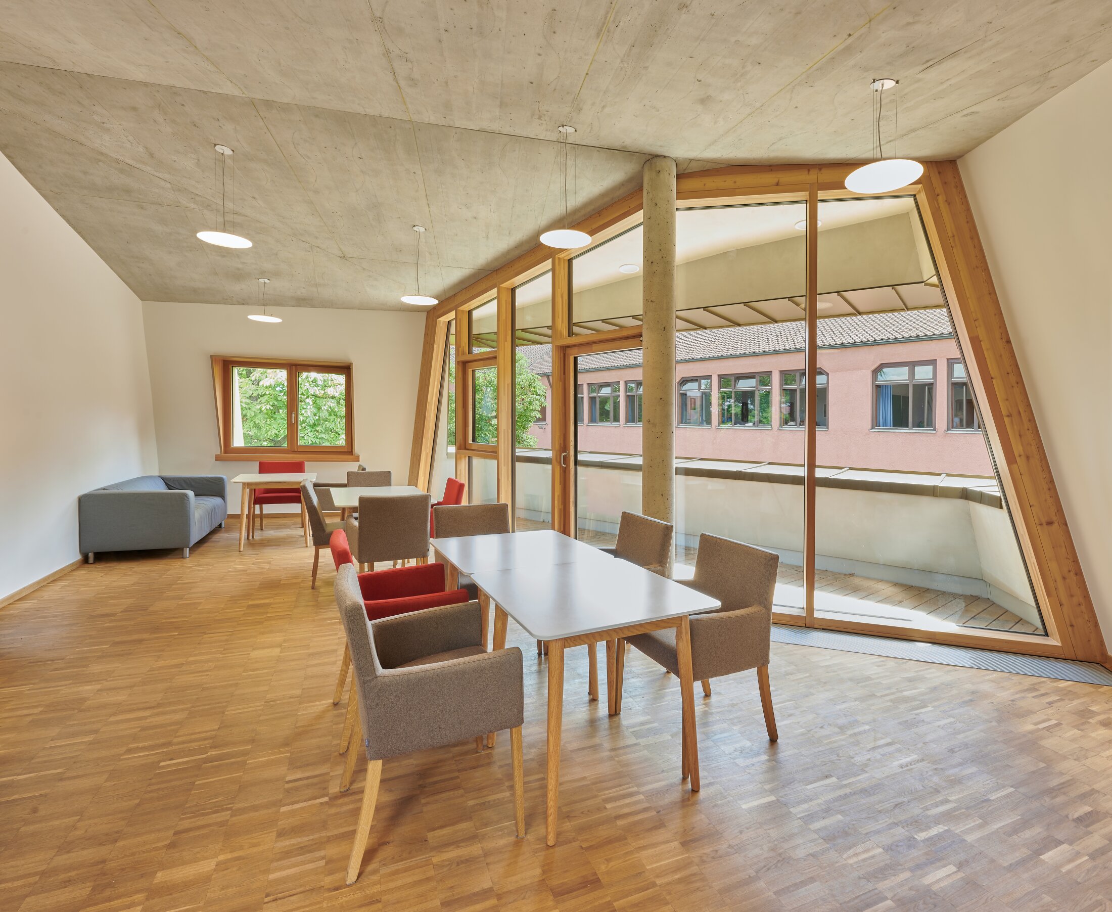 Project by Behnisch Architekturbuero, Freie Waldorfschule Uhlandshoehe - New School and Administration Building. Bright room with tables and chairs, large slanted windows, wooden floor and a sofa in the corner. The windows overlook the balcony.