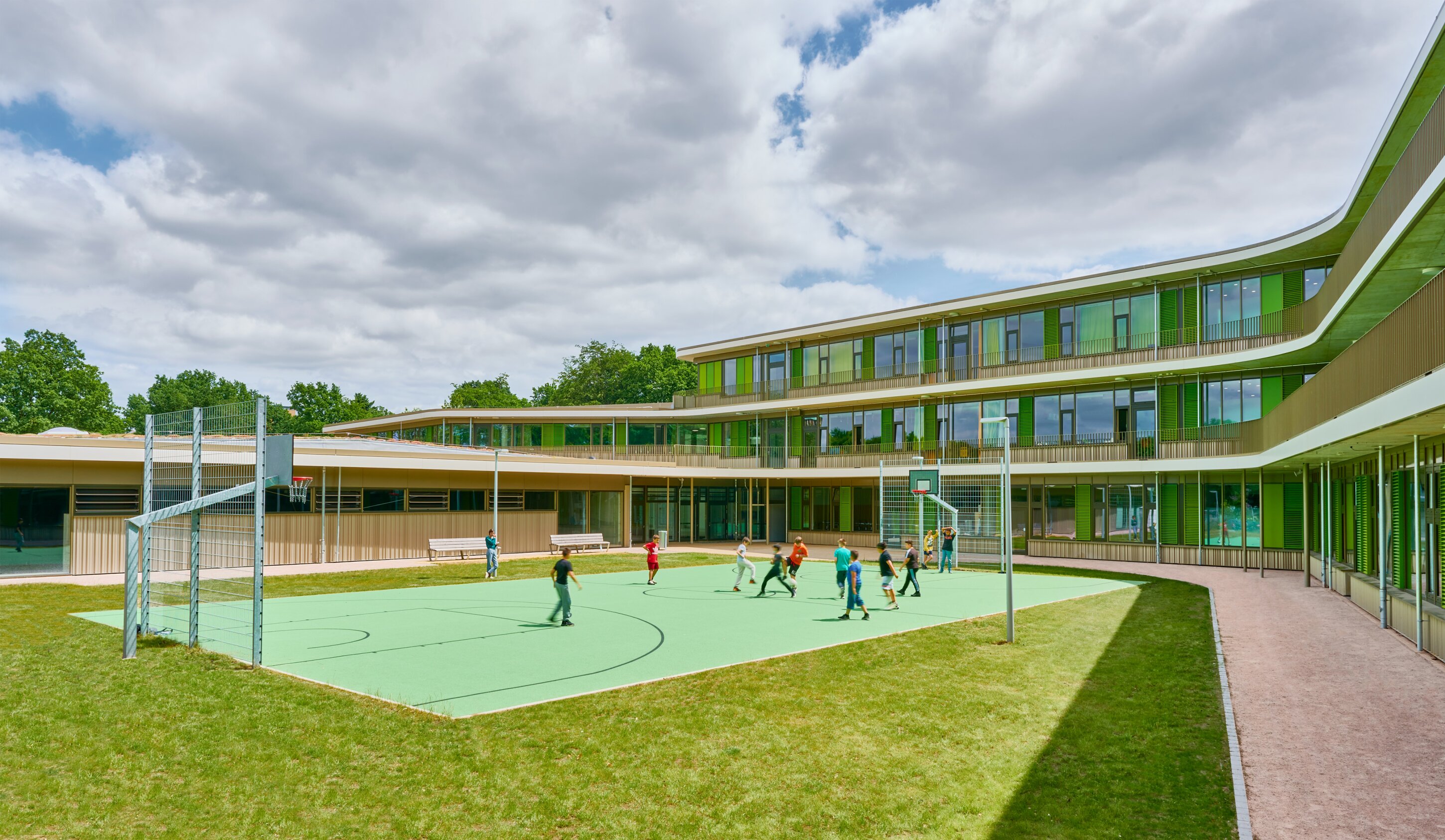 Project by Behnisch Architekturbuero, Lurup Neighborhood School. School courtyard with a basketball court where children are playing, surrounded by a modern building with green accents and large windows.