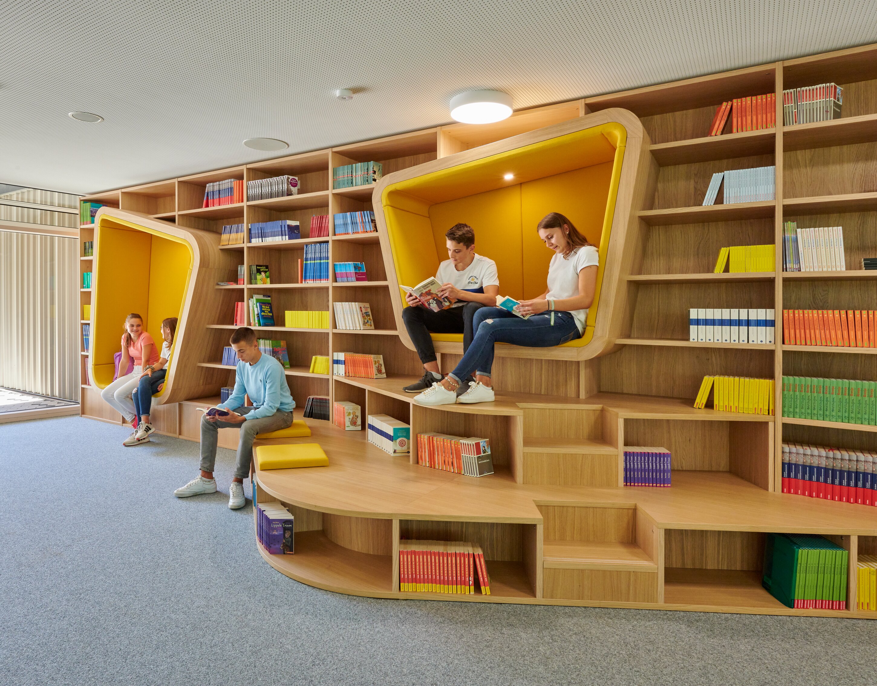 Project by Behnisch Architekturbuero, Lurup Neighborhood School. Library area with wooden shelves and built-in yellow reading nooks, where several students sit and read books.