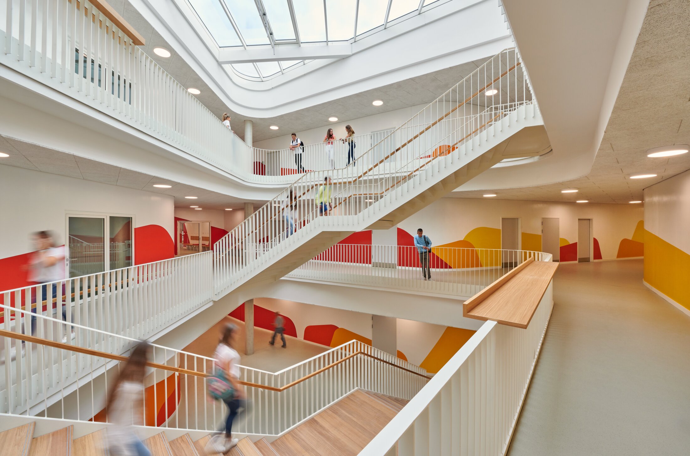 Project by Behnisch Architekturbuero, Lurup Neighborhood School. Multi-level school interior with open atrium, white railings and stairs, students moving between floors, and colorful wall graphics.