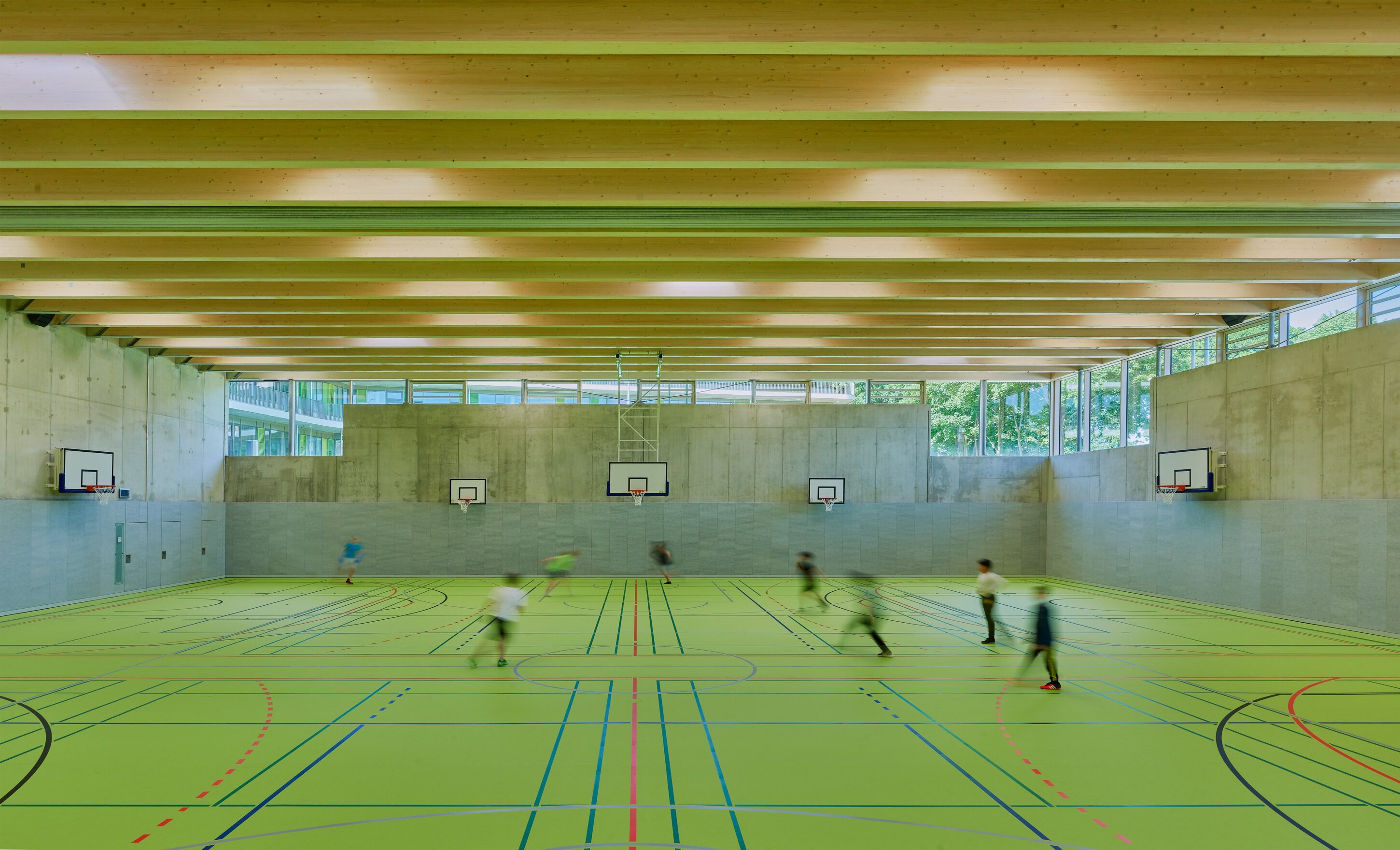 Project by Behnisch Architekturbuero, Lurup Neighborhood School. Indoor gym with green sports floor and multiple basketball hoops, where several people are moving across the court.