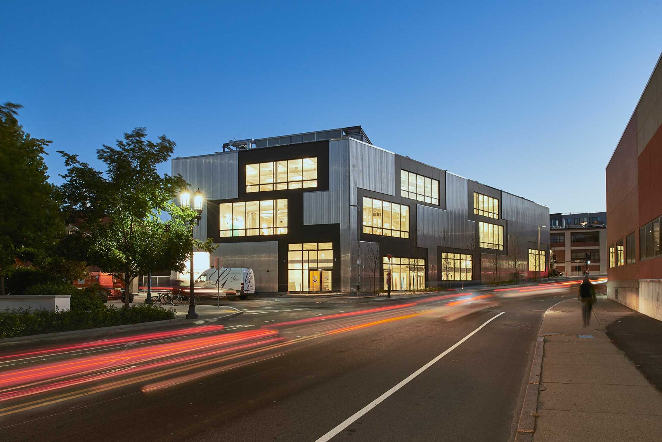 Project by Behnisch Architekturbuero, Artists for Humanity EpiCenter Expansion. Modern building with illuminated windows at dusk along a street, with blurred car light trails and a pedestrian on the sidewalk.