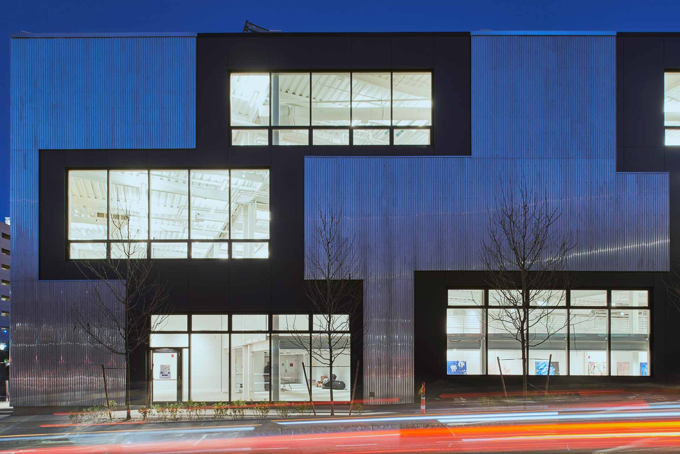 Project by Behnisch Architekturbuero, Artists for Humanity EpiCenter Expansion. Modern building facade at night with large lit windows, corrugated metal panels, and blurred light trails from passing cars in front.
