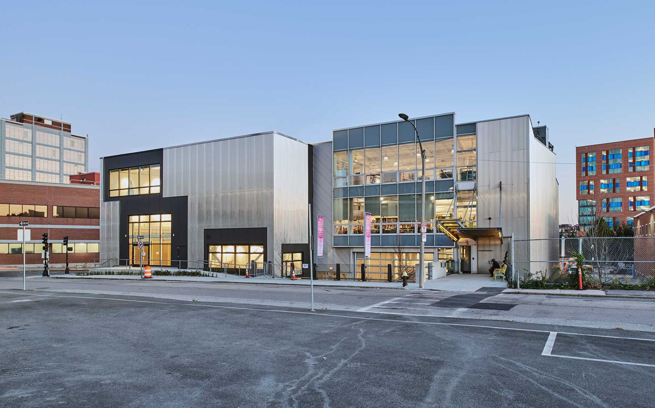 Project by Behnisch Architekturbuero, Artists for Humanity EpiCenter Expansion. Modern multi-story building with metal and glass facade, large windows, and street intersection in foreground at dusk.