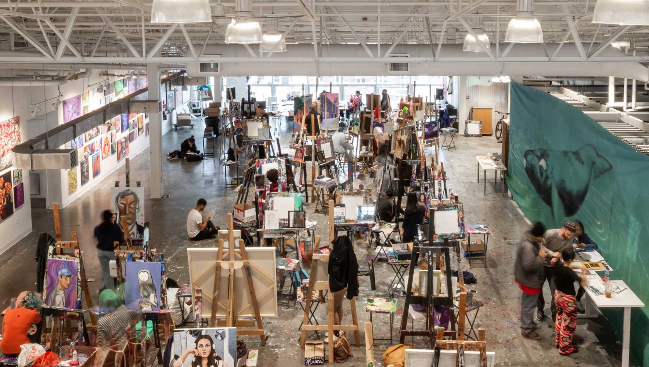 Project by Behnisch Architekturbuero, Artists for Humanity EpiCenter Expansion. Large open art studio with many people painting at easels, artworks on walls, and tables with supplies under bright industrial lighting.