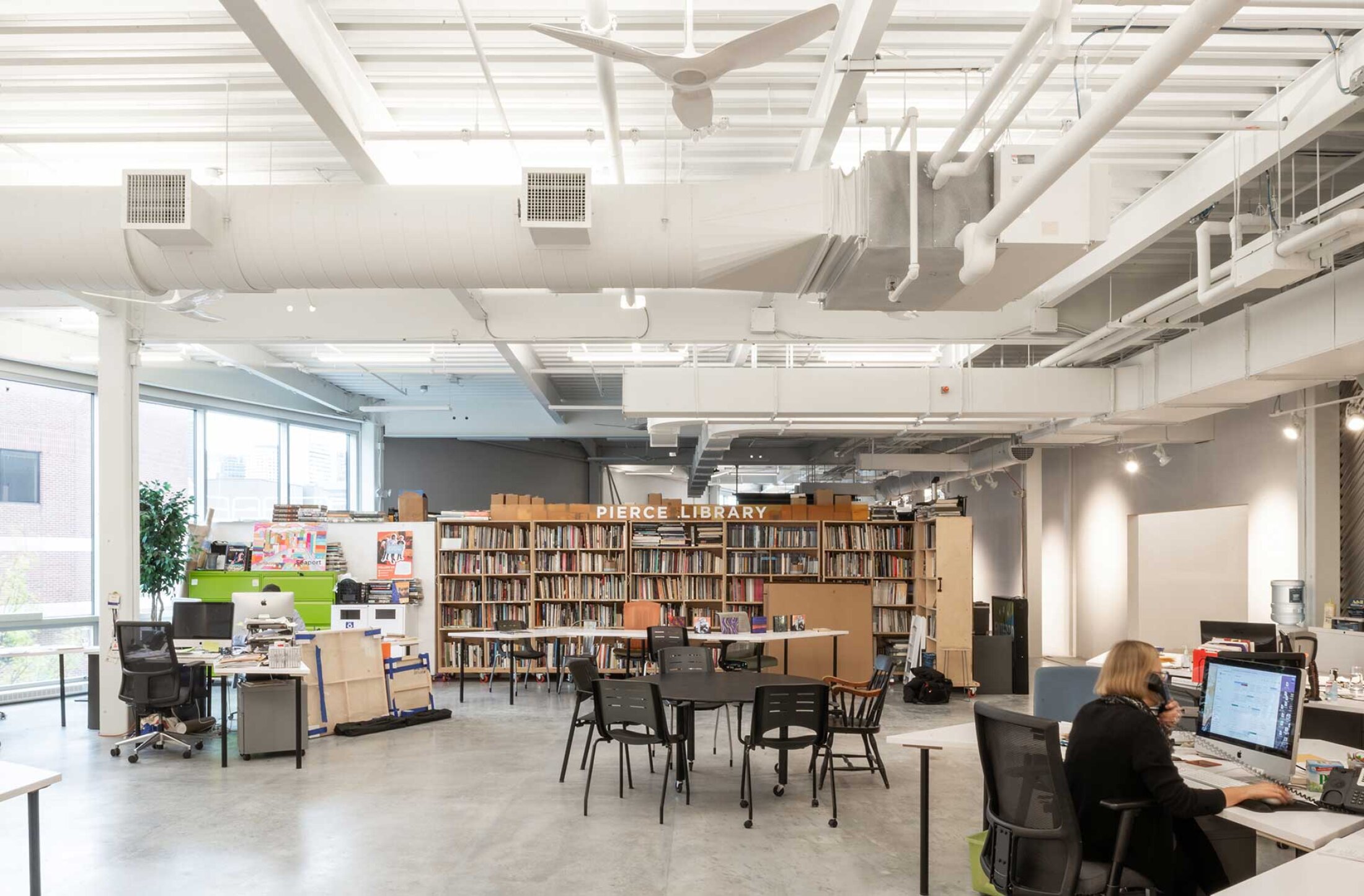 Project by Behnisch Architekturbuero, Artists for Humanity EpiCenter Expansion. Open library workspace with bookshelves, tables and chairs, desks with computers, and exposed ceiling ducts under bright lighting.