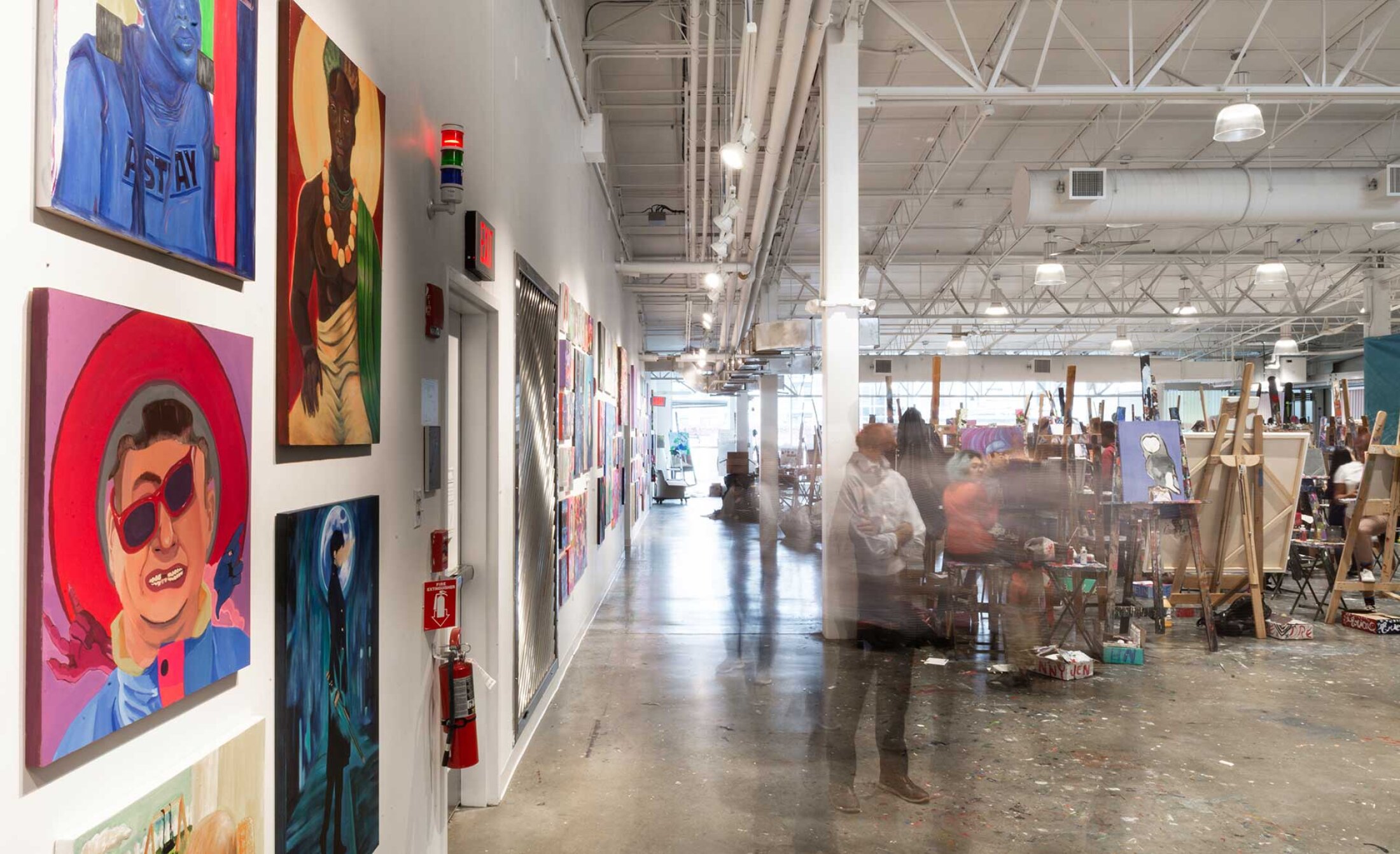 Project by Behnisch Architekturbuero, Artists for Humanity EpiCenter Expansion. Art studio interior with paintings displayed on a wall, people working at easels, and motion blur of figures moving through the space.