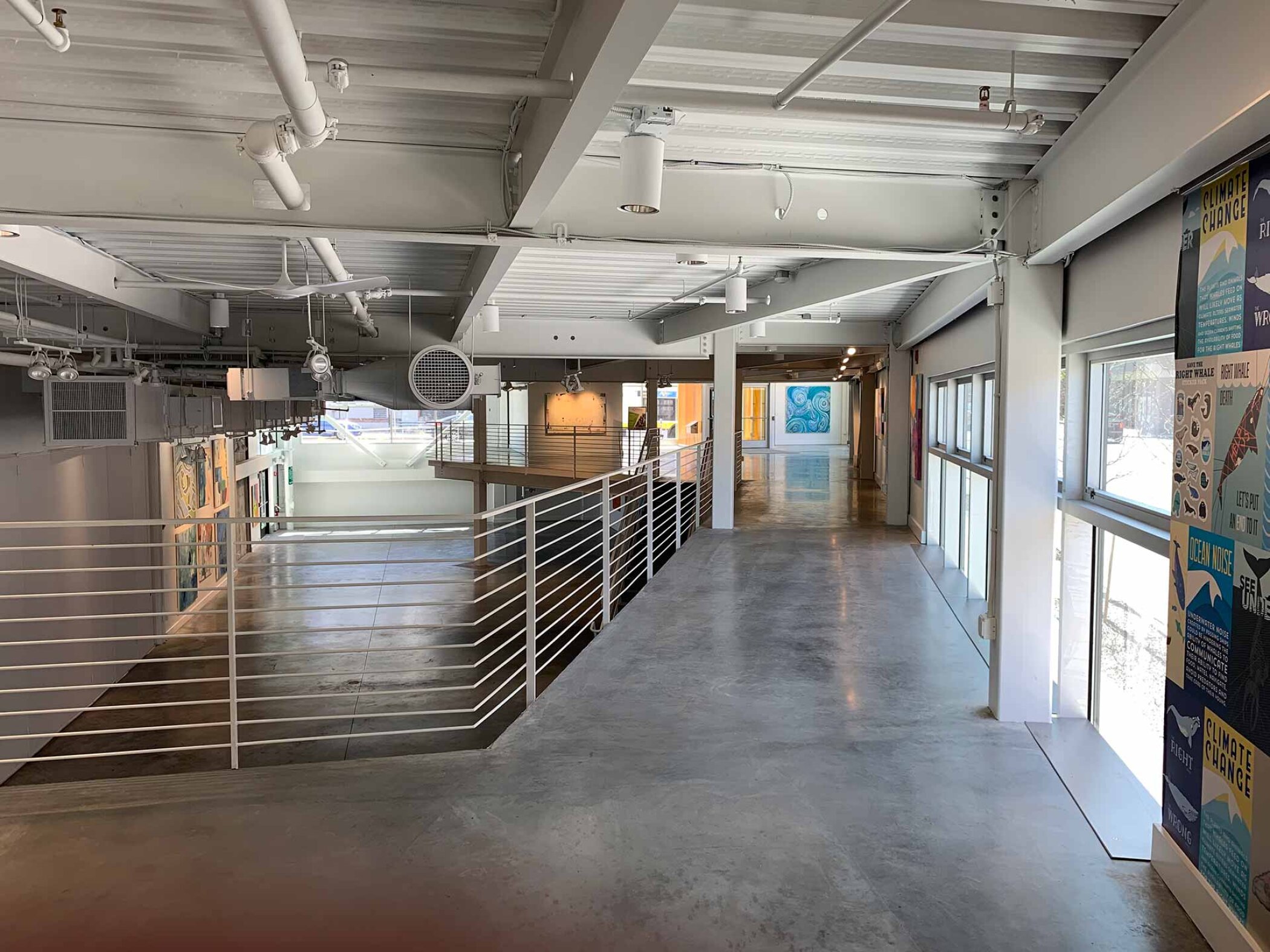 Project by Behnisch Architekturbuero, Artists for Humanity EpiCenter Expansion. Open interior corridor with metal railing, exposed ceiling ducts, large windows, and artwork displayed along the walls.