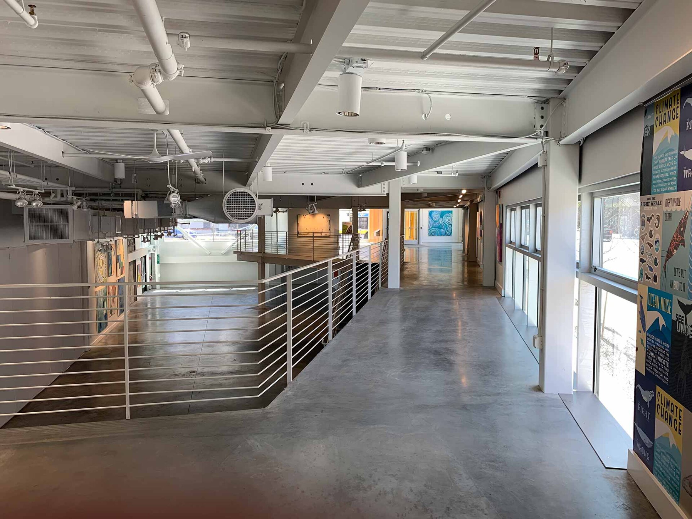 Project by Behnisch Architekturbuero, Artists for Humanity EpiCenter Expansion. Open interior corridor with metal railing, exposed ceiling ducts, large windows, and artwork displayed along the walls.