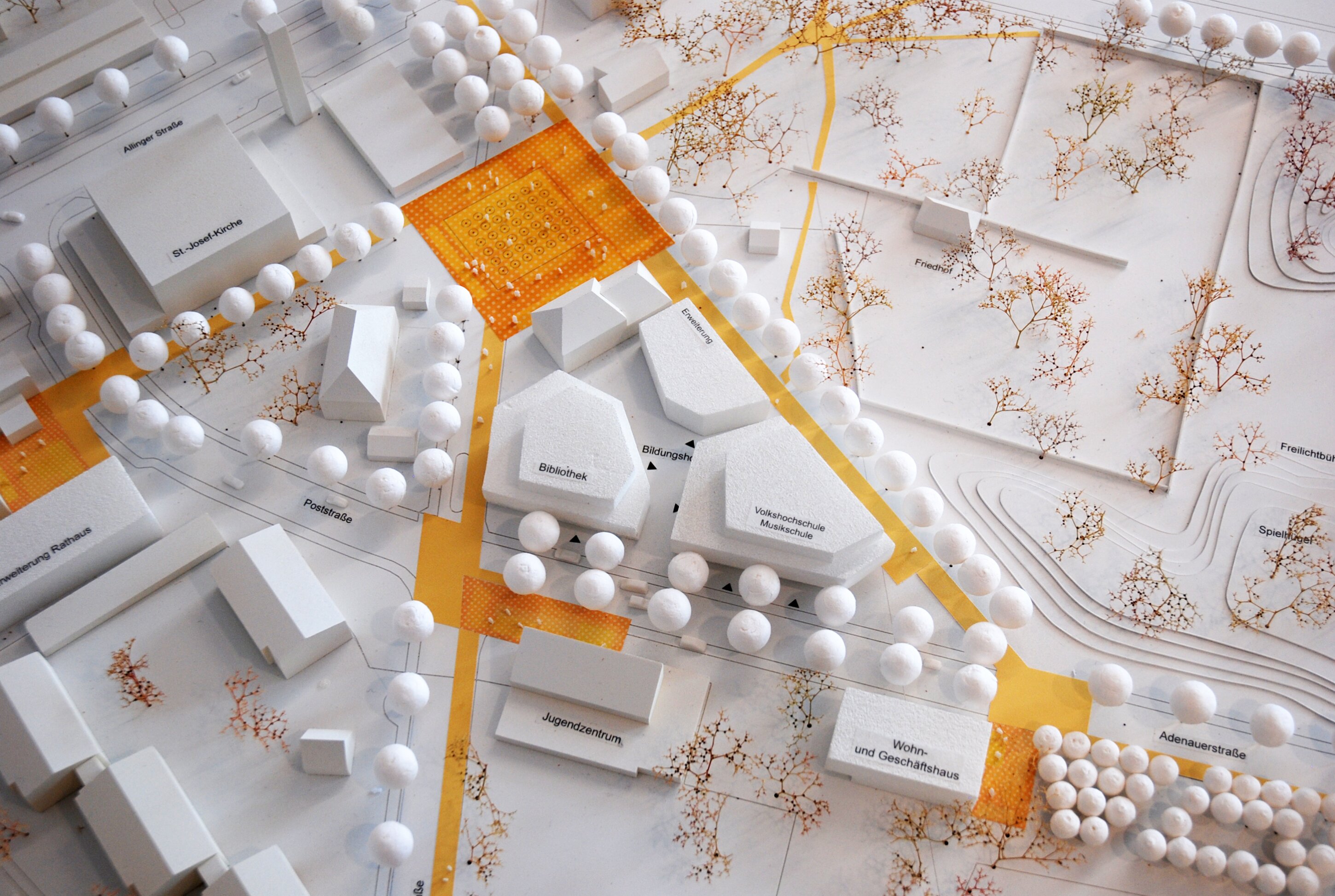 Project by Behnisch Architekturbuero, City Center Development Puchheim. Top view of an urban planning model with labeled buildings, streets, trees, and highlighted paths in orange across a neighborhood layout.