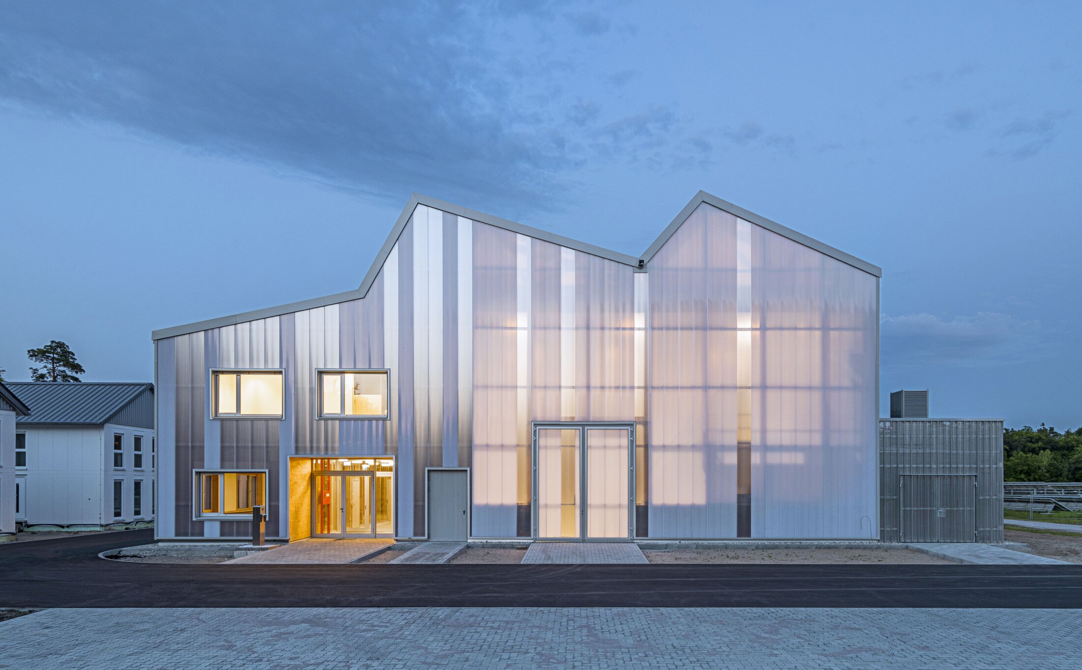 Project by Behnisch Architekturbuero, KIT Energy Lab 2.0. Modern building with a polycarbonate envelope that glows at dusk. The building is formed with sloping roof shapes and an illuminated entrance on a street.