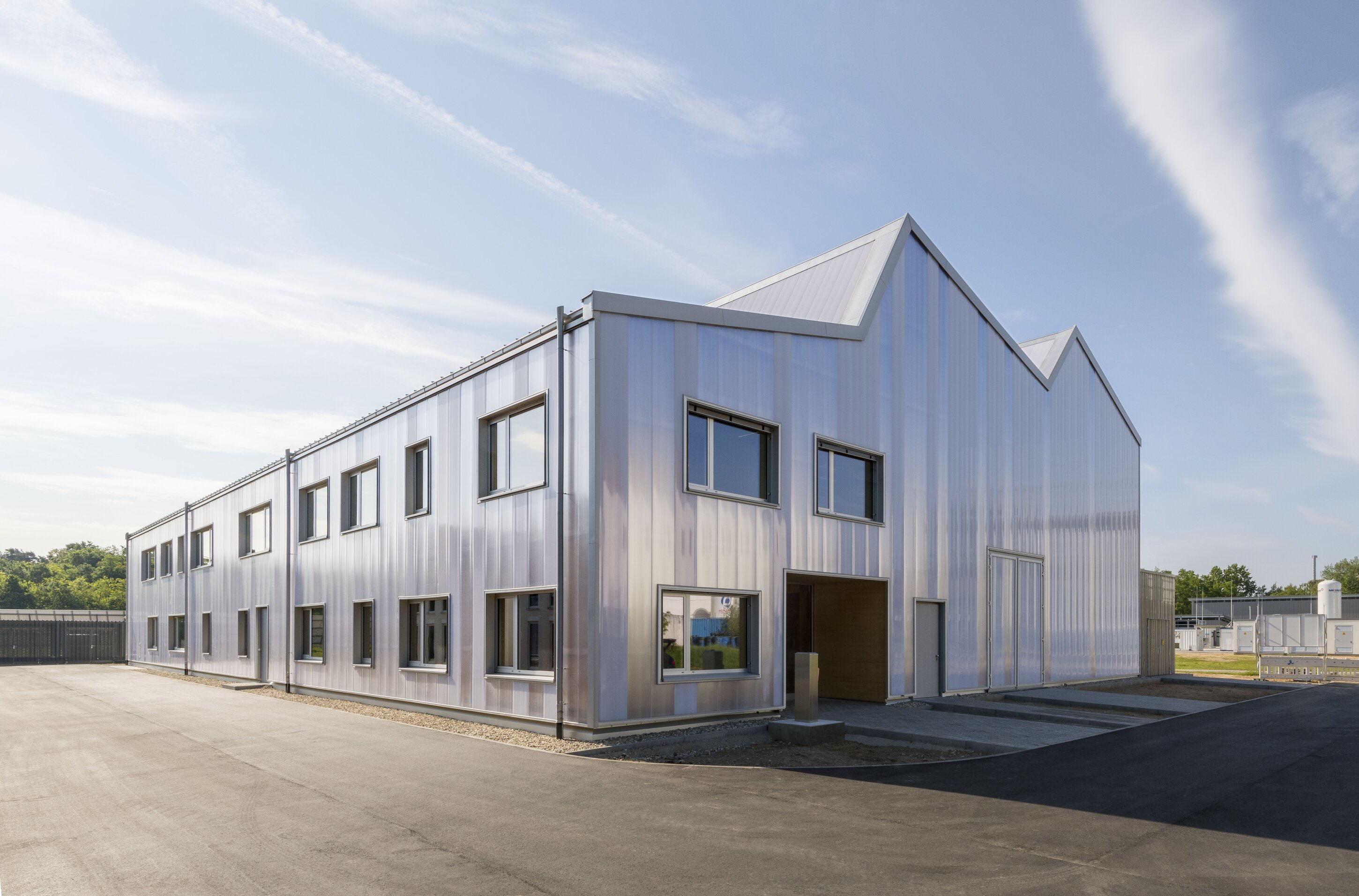 Project by Behnisch Architekturbuero, KIT Energy Lab 2.0. Industrial-style building with translucent facade and sawtooth roof, multiple windows, and paved surroundings under a partly cloudy sky.