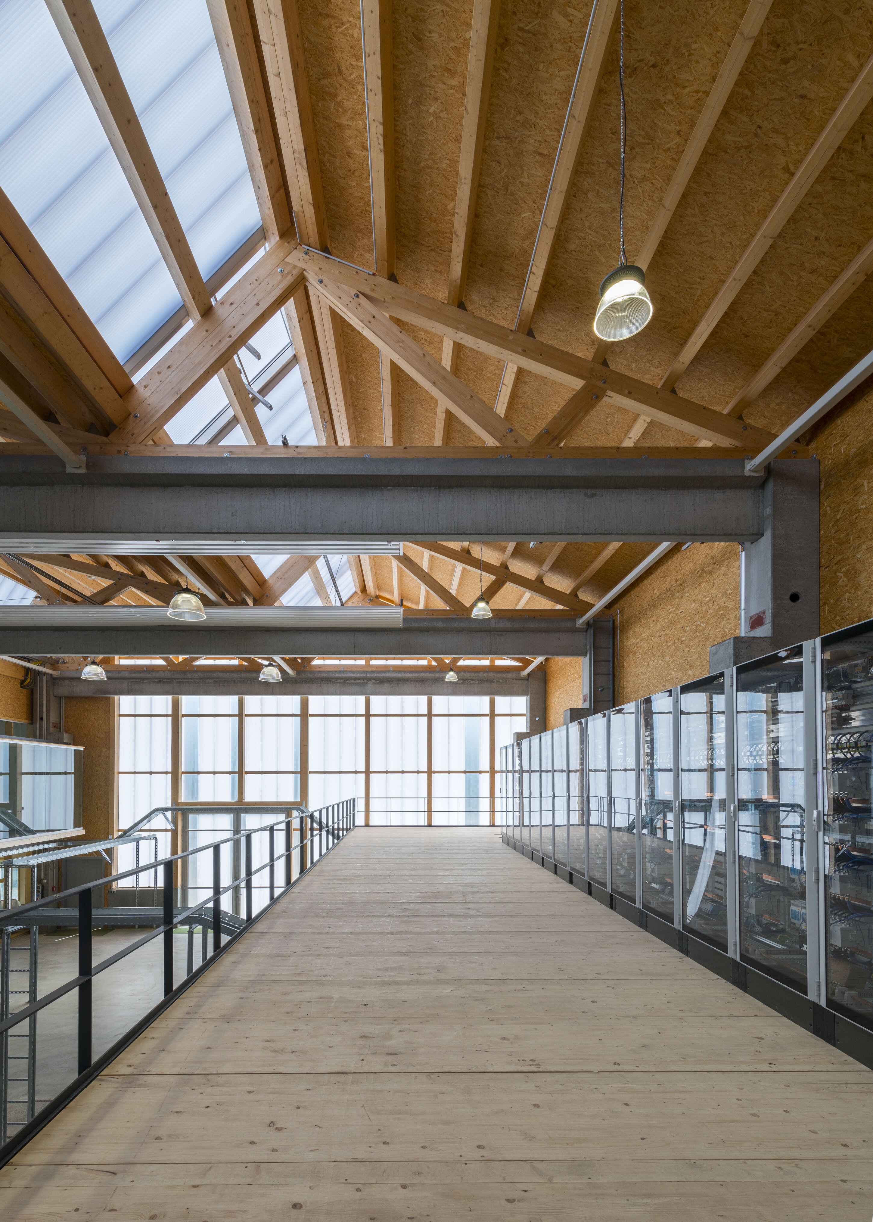Project by Behnisch Architekturbuero, KIT Energy Lab 2.0. Upper-level walkway in a modern interior with timber roof trusses, skylights, glass railings, and rooms enclosed by glass partitions.