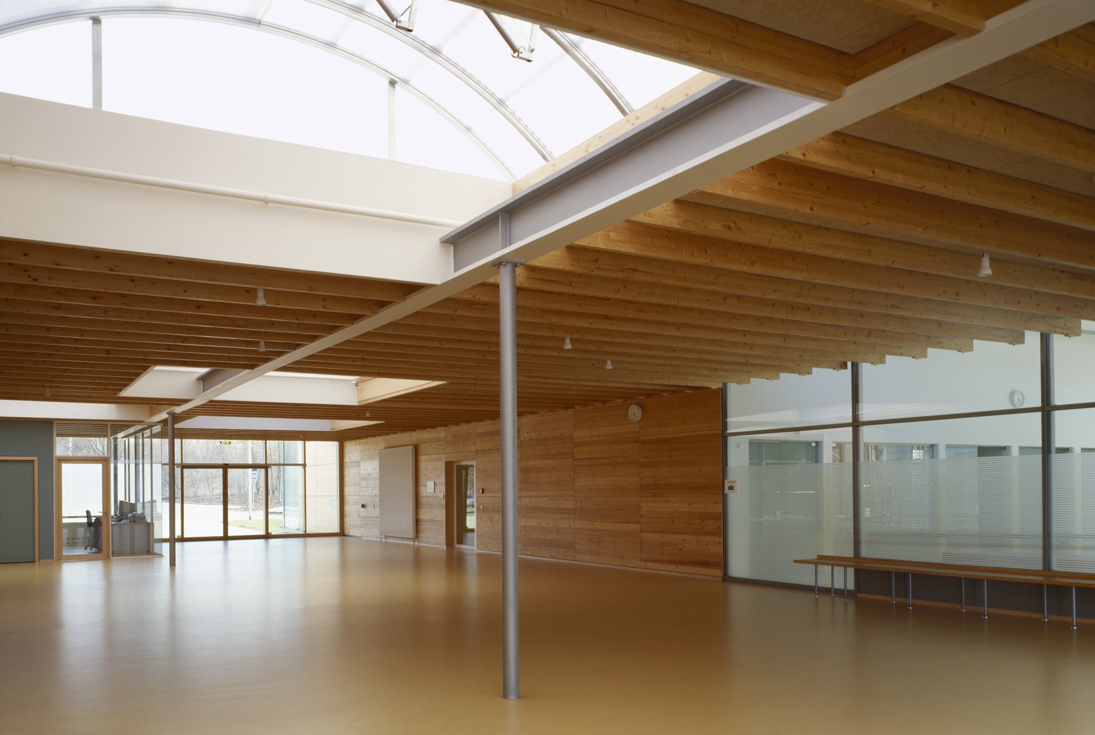 Project by Behnisch Architekturbuero, Pistorius School Herbrechtingen. Spacious interior hall with wood-paneled walls and ceiling, skylights above, glass partitions, and a bench along one wall.