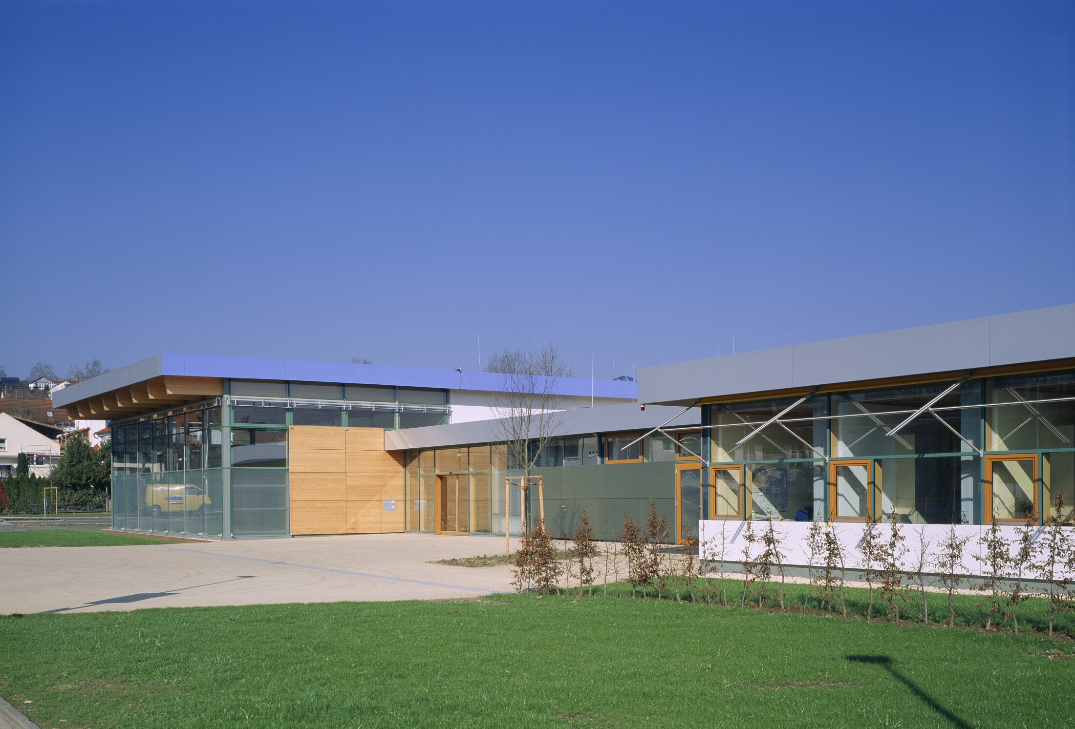 Project by Behnisch Architekturbuero, Pistorius School Herbrechtingen. Low school building with glass and wood facade, next to a paved square and a green lawn under a clear blue sky.