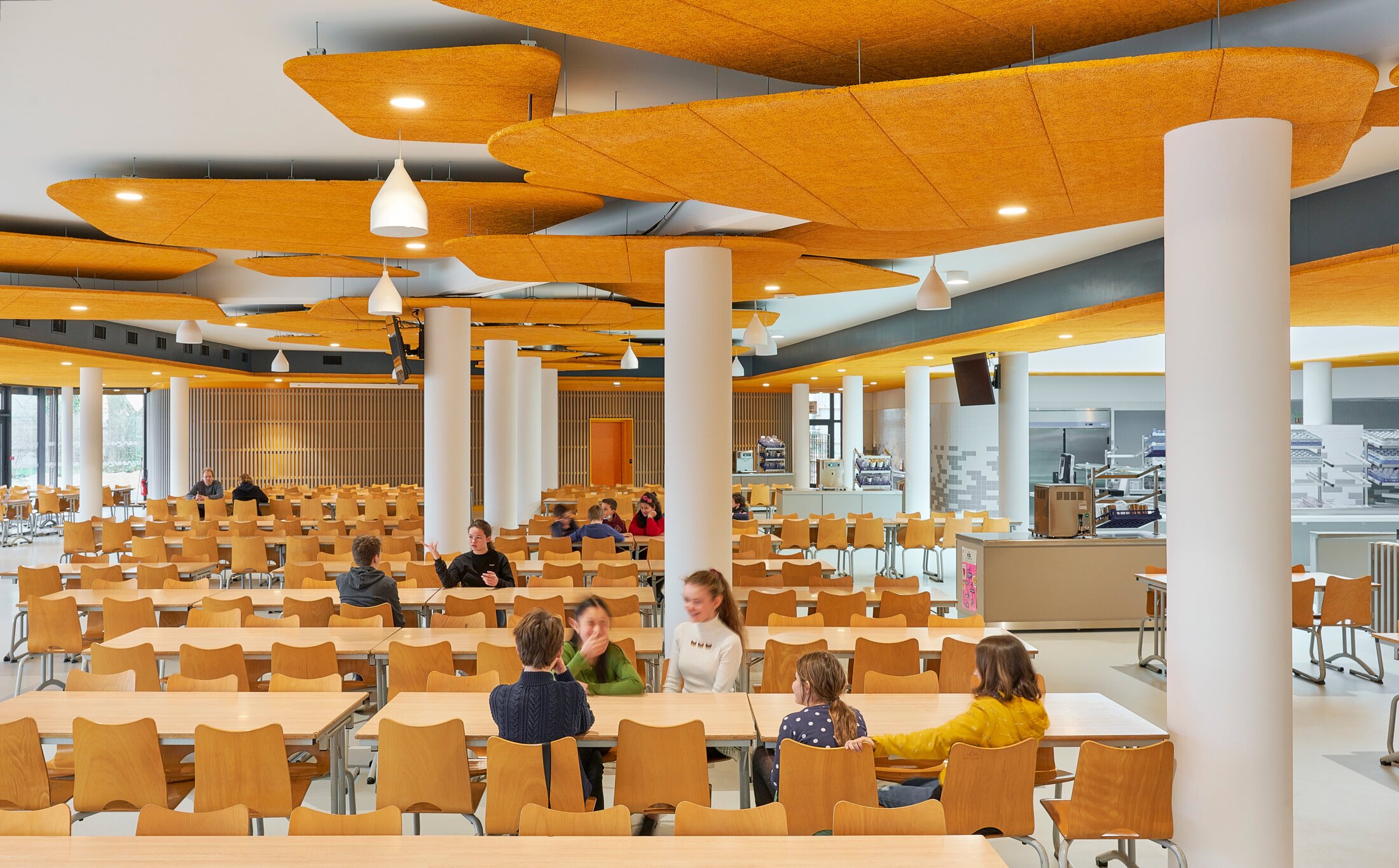 Project by Behnisch Architekturbuero, Lycee Franco Allemand. School cafeteria with rows of tables and chairs, children seated and talking, orange acoustic ceiling panels and serving area in background.
