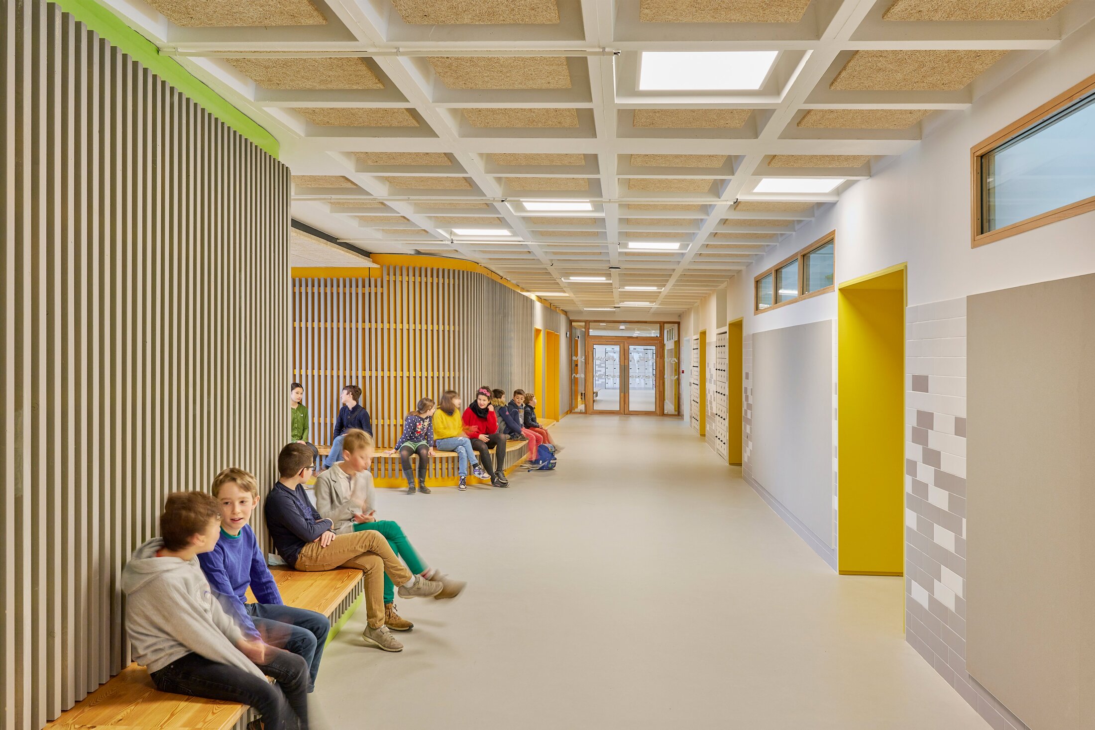 Project by Behnisch Architekturbuero, Lycee Franco Allemand. School corridor with children sitting on wooden benches along a wood-paneled wall; bright yellow doors and lockers line the corridor.