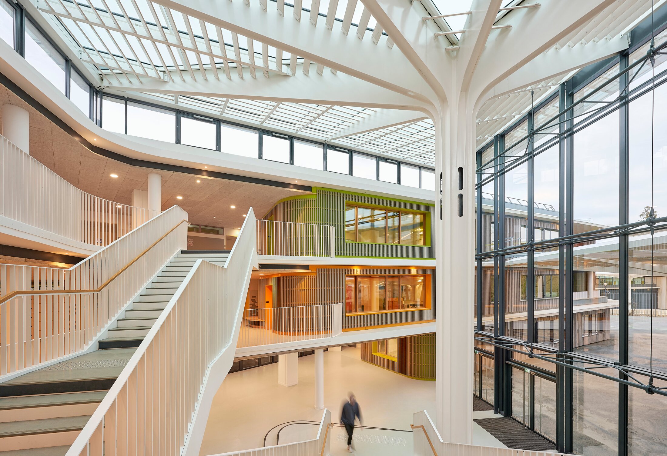 Project by Behnisch Architekturbuero, Lycee Franco Allemand. A bright, multi-story atrium with white stairwells, large glass walls and a central column, featuring open balconies and interior windows. Each of the three floors is distinguished by a color accent.