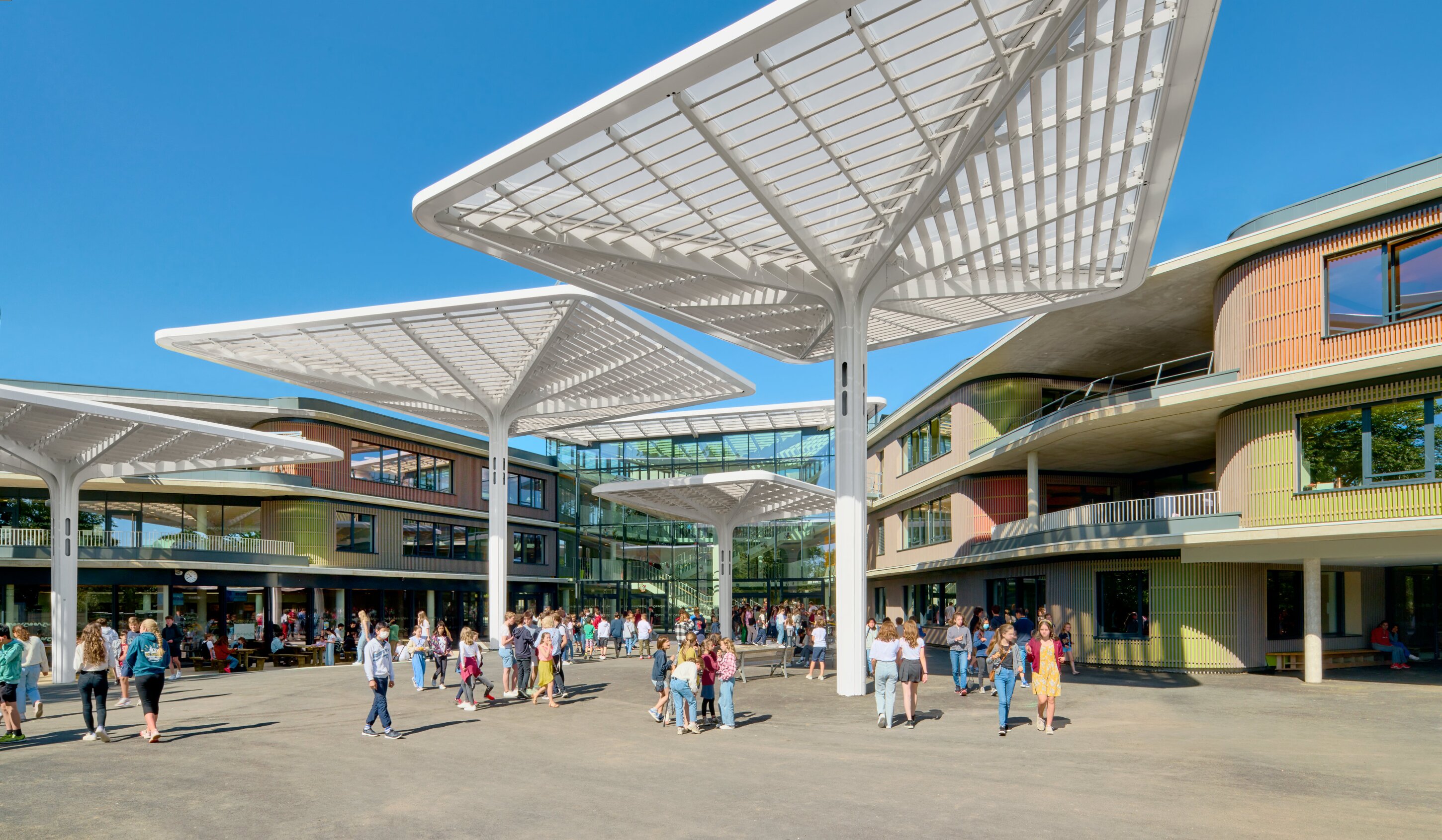 Project by Behnisch Architekturbuero, Lycee Franco Allemand. School courtyard with many students gathered under large white canopy structures between modern buildings.