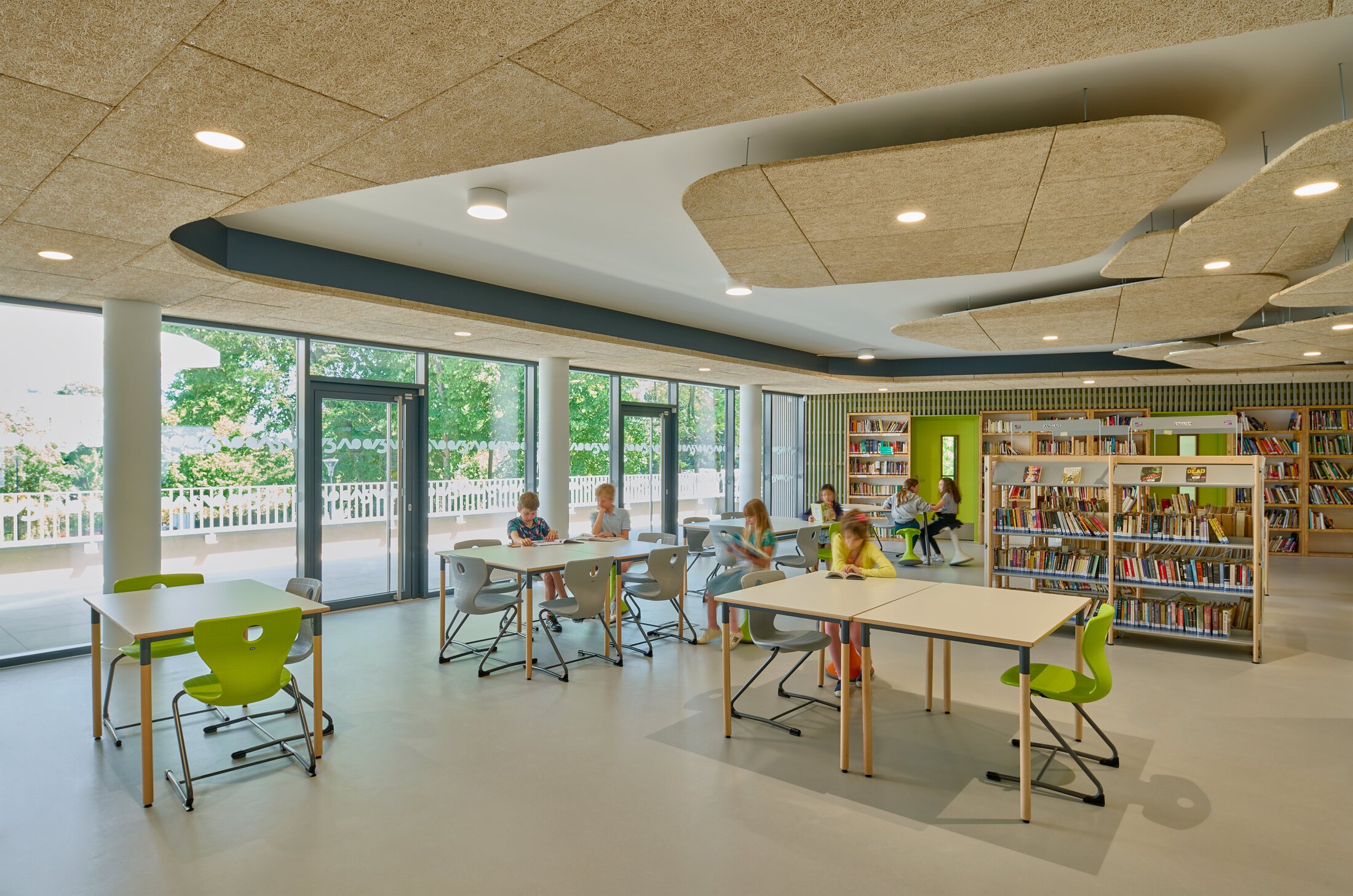 Project by Behnisch Architekturbuero, Lycee Franco Allemand. Library classroom with children reading at tables, bookshelves along the wall, and large windows opening to an outdoor terrace.