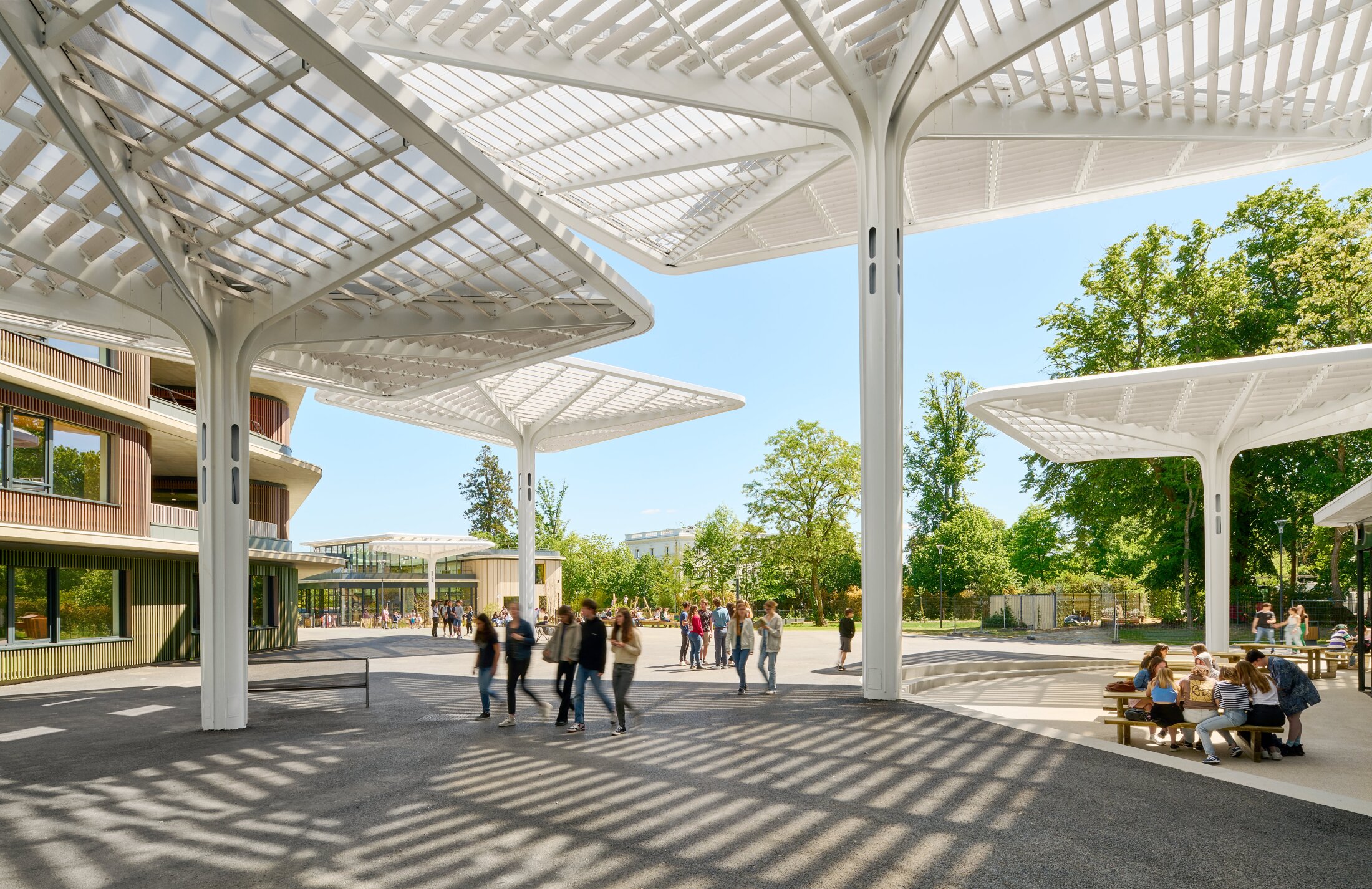 Project by Behnisch Architekturbuero, Lycee Franco Allemand. Outdoor plaza with large white canopy structures, people walking and sitting, trees and buildings surrounding the open public space.