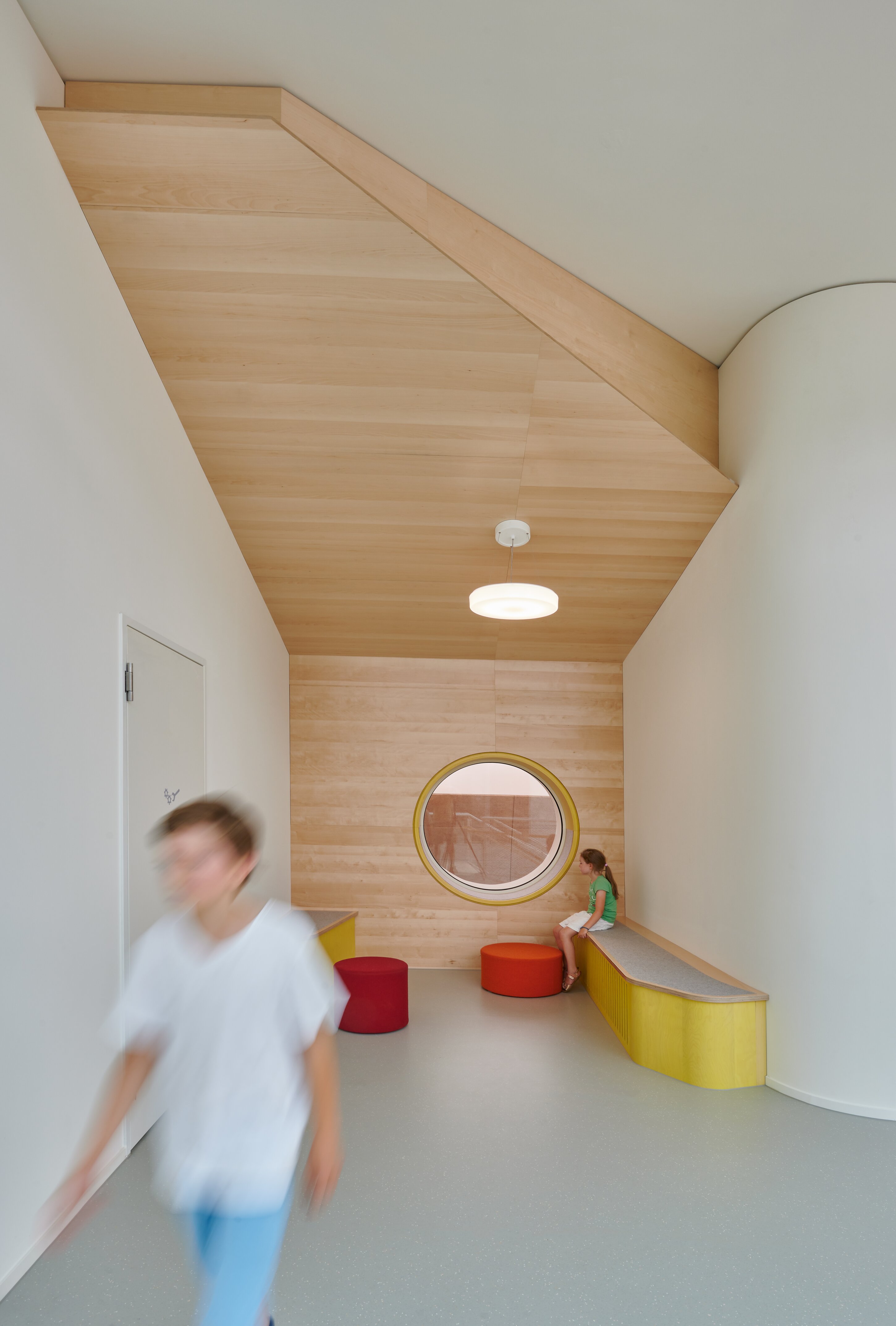 Project by Behnisch Architekturbuero, Ludwig-Weber School. A narrow school hallway with a wooden ceiling, a round window, and a built-in bench; one child is sitting, another is walking past. The seated child is sitting on a yellow cushion. There are also red stools.