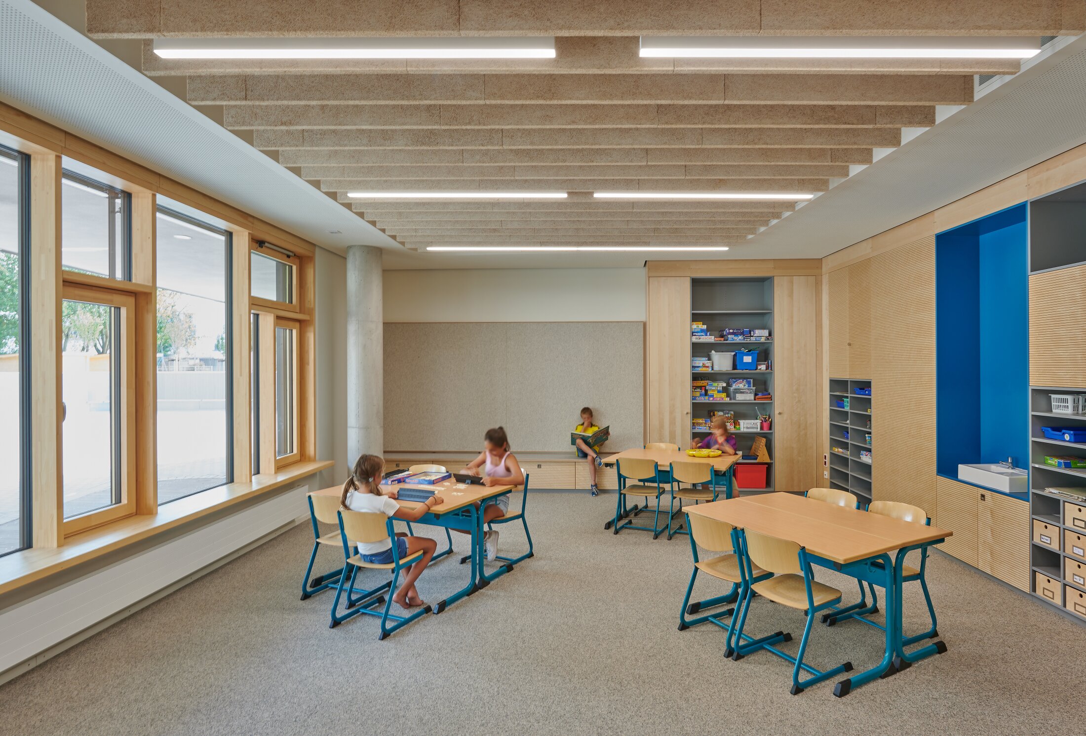 Project by Behnisch Architekturbuero, Gotthard-M&uuml;ller-School Bernhausen. Classroom with tables and chairs, children reading and playing, storage shelves on wall and large windows along one side.