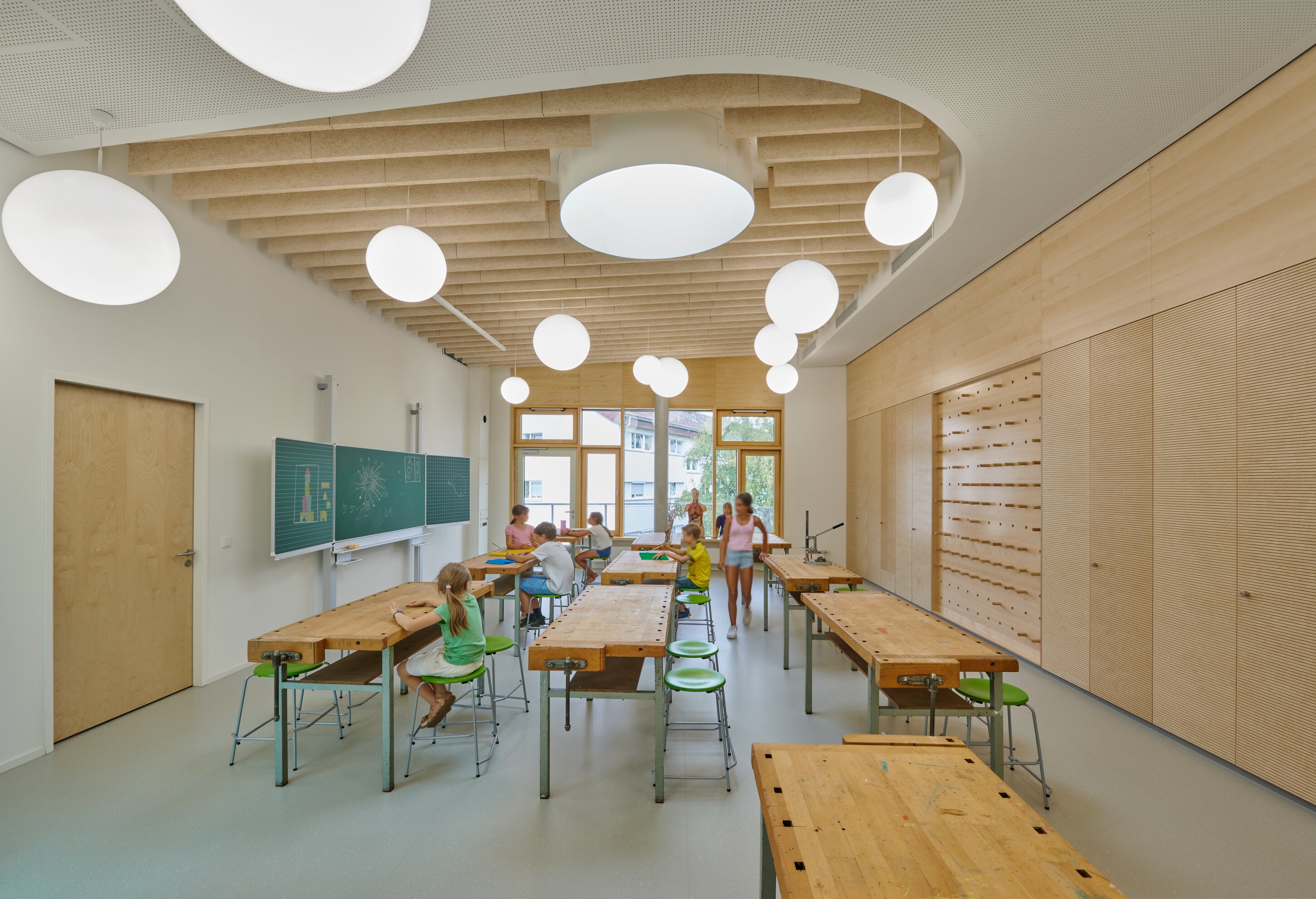 Project by Behnisch Architekturbuero, Ludwig-Weber School. Classroom workshop with wooden tables and stools, children working, chalkboards on wall, large windows and round ceiling lights.