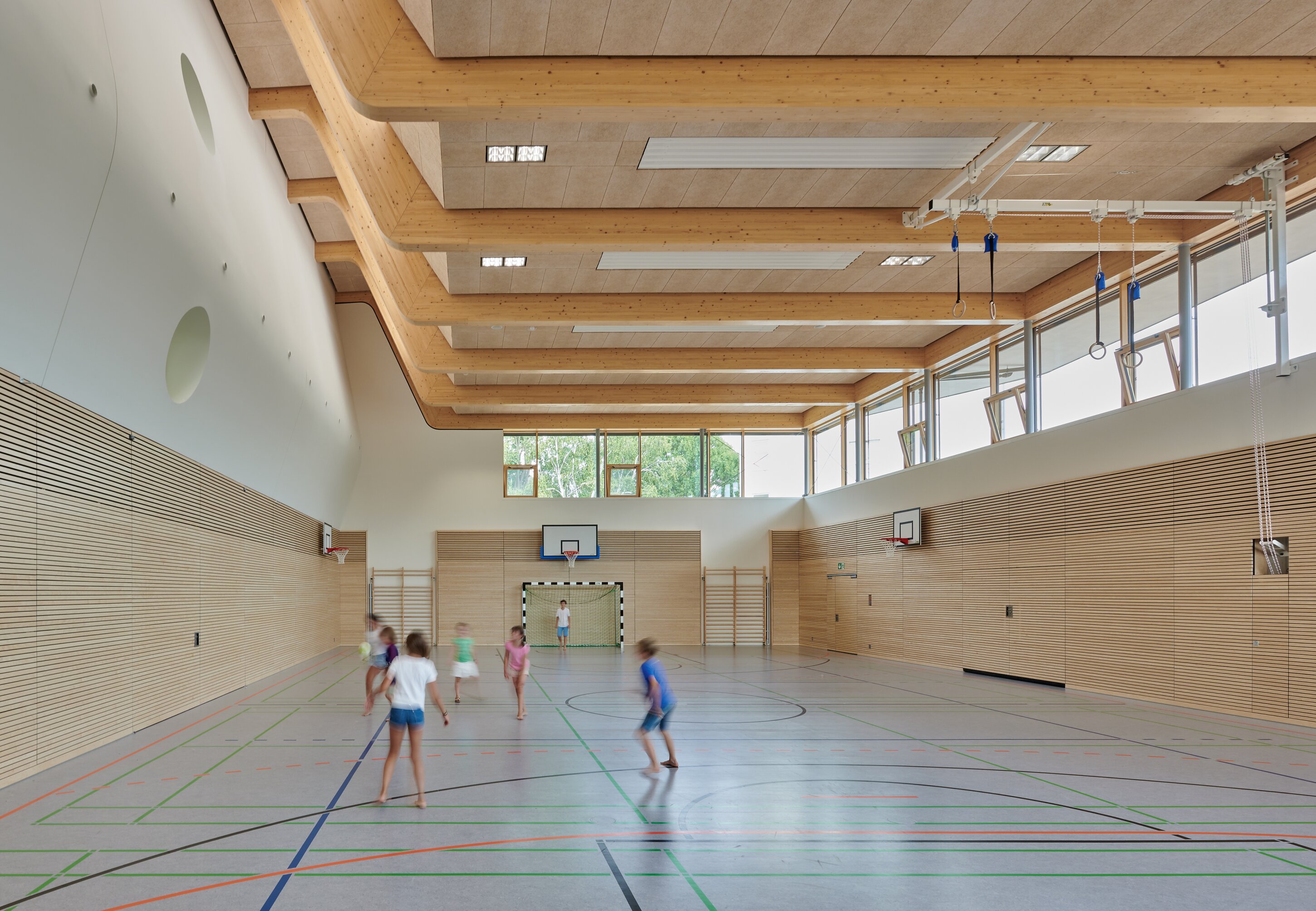 Project by Behnisch Architekturbuero, Ludwig-Weber School. School gym with wooden walls and high ceiling, children running and playing on marked court, basketball hoops and goal at far end.