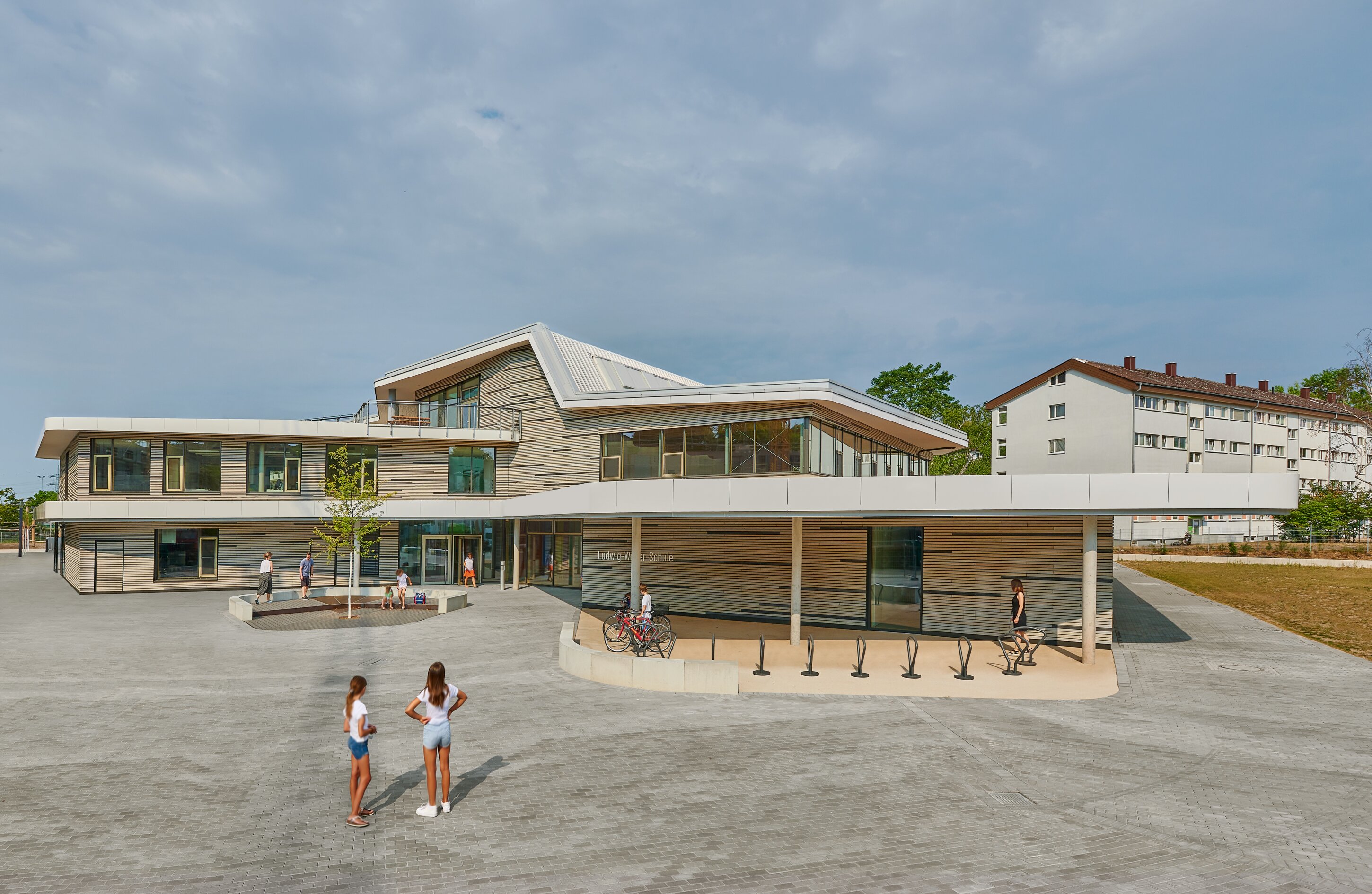 Project by Behnisch Architekturbuero, Ludwig-Weber School. A modern school building with an angular roof and glass facade; students stand and walk in a large paved courtyard. There are several bicycle parking areas in front of the building.