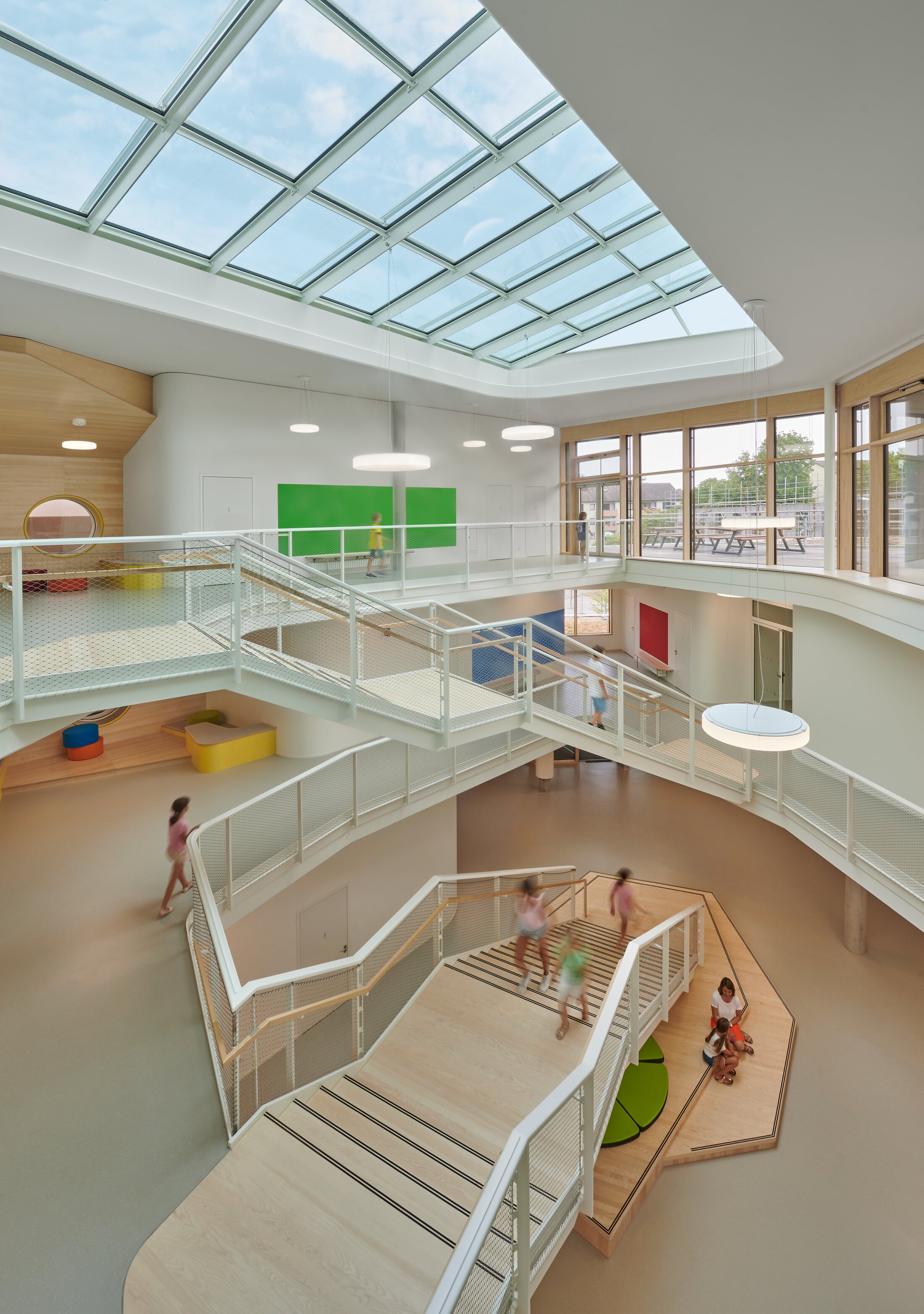 Gotthard-M&uuml;ller-School Bernhausen. A multi-story school atrium with a glass roof, staircases and corridors, children moving between the levels, and open learning areas. There are occasional splashes of color within the atrium, such as green, yellow, red, and blue.