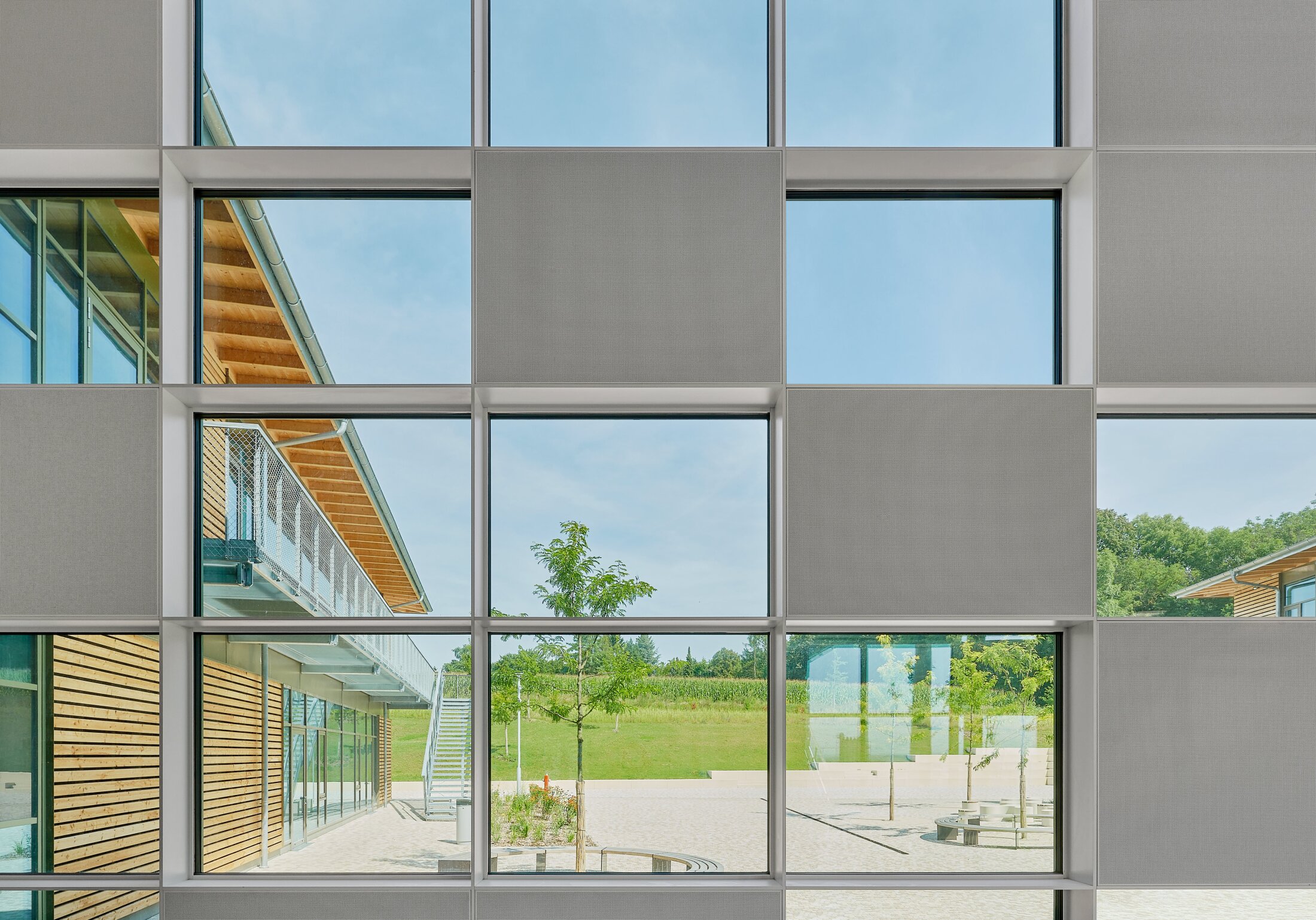 Project by Behnisch Architekturbuero, Public Secondary School Neuburg an der Donau. Grid of large windows with opaque panels, showing view of the school courtyard with trees, benches, and a wood-clad building with exterior stairs.