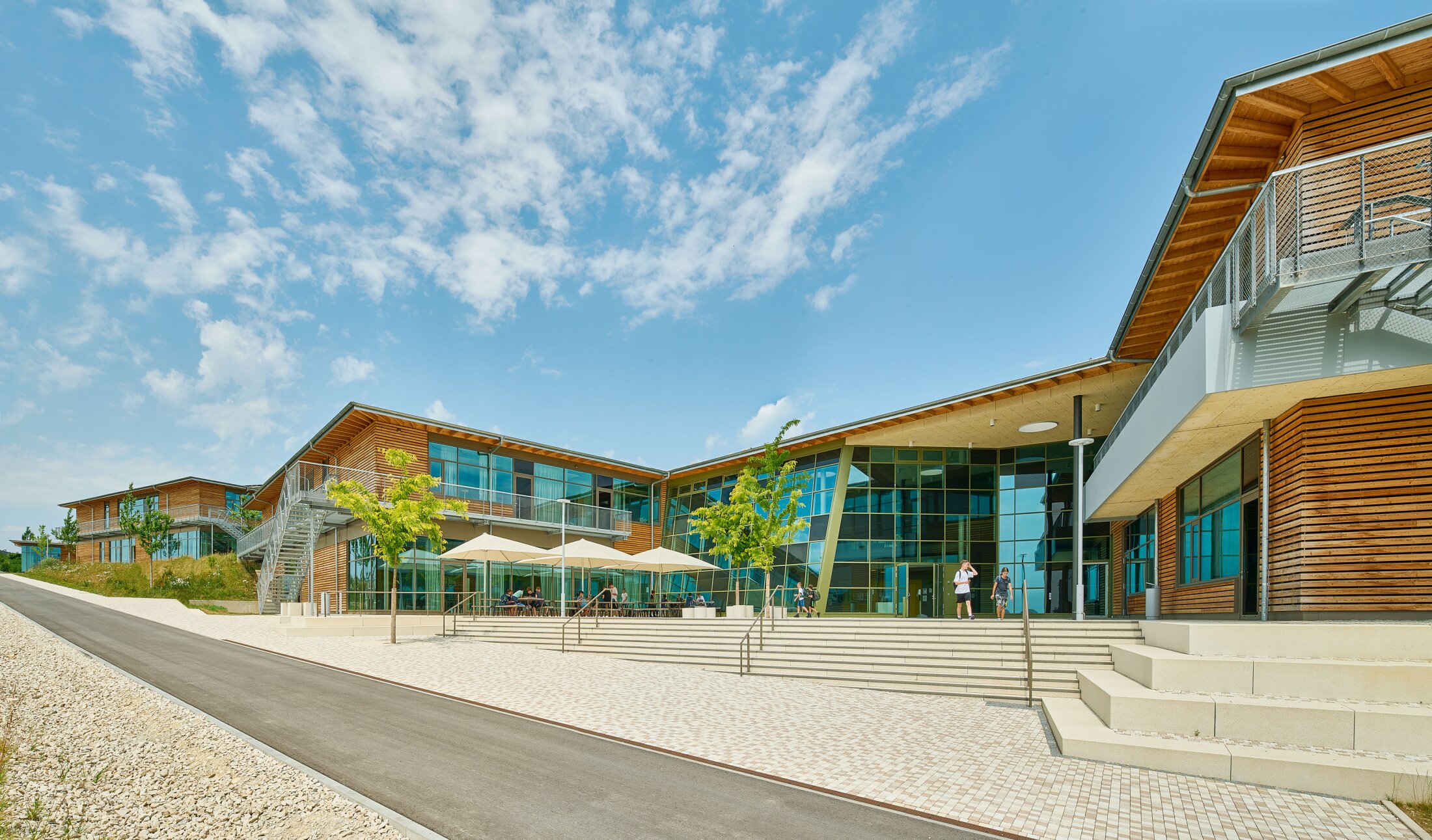 Project by Behnisch Architekturbuero, Public Secondary School Neuburg an der Donau. A modern, single-story building with wood cladding and a large glass facade, featuring outdoor seating with parasols. The building is accessed via spacious external staircases. Several students are standing in front of the entrance.