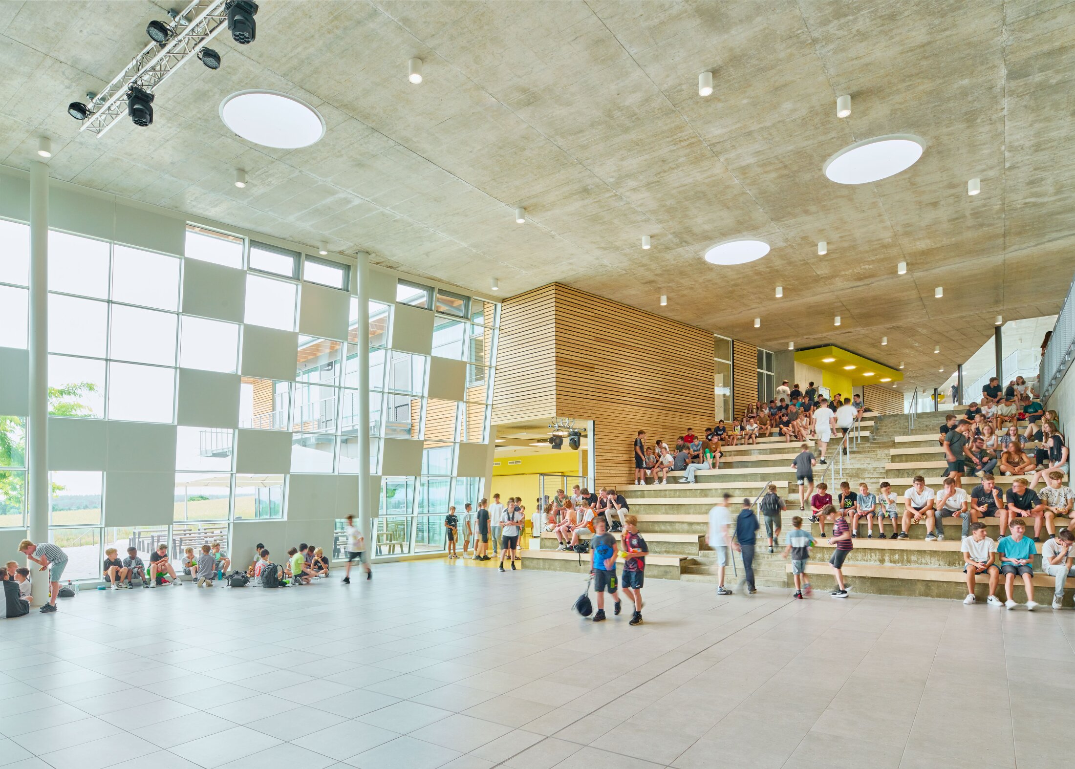 Project by Behnisch Architekturbuero, Public Secondary School Neuburg an der Donau. Spacious school atrium with large windows, open floor, and tiered steps where many students sit and stand.