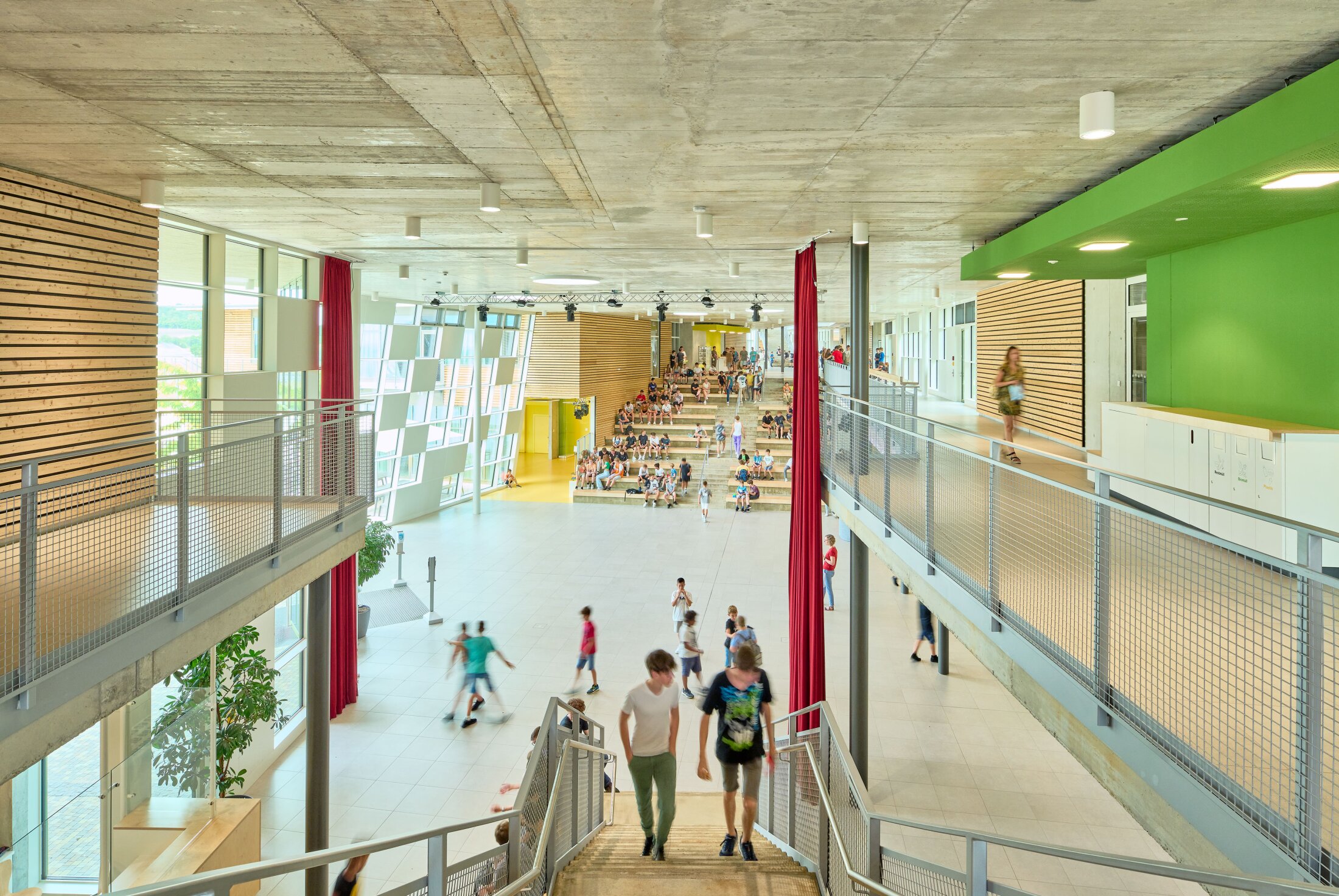 Project by Behnisch Architekturbuero, Public Secondary School Neuburg an der Donau. Large school atrium with stairs, balconies, and red curtains; many students walk and sit on stepped seating in the background.