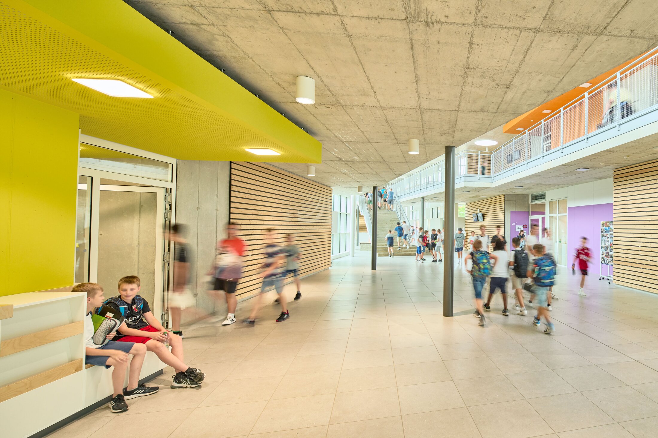 Project by Behnisch Architekturbuero, Public Secondary School Neuburg an der Donau. School corridor with students walking and sitting, yellow ceiling feature, wood wall panels, and stairs leading to an upper-level walkway.