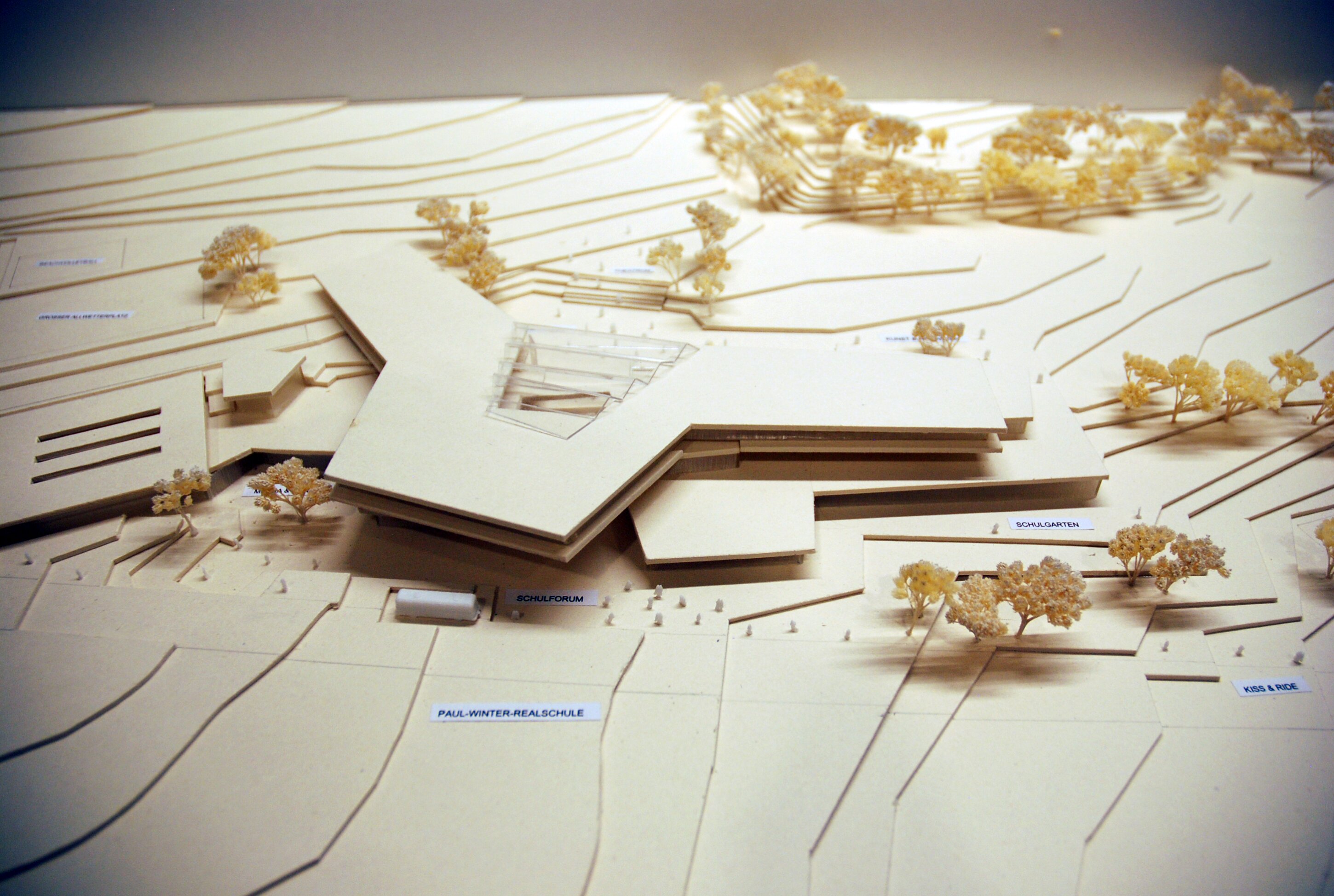 Project by Behnisch Architekturbuero, Public Secondary School Neuburg an der Donau. Architectural model of a school complex on a contoured site, with layered terrain, buildings, and small trees arranged across the landscape.