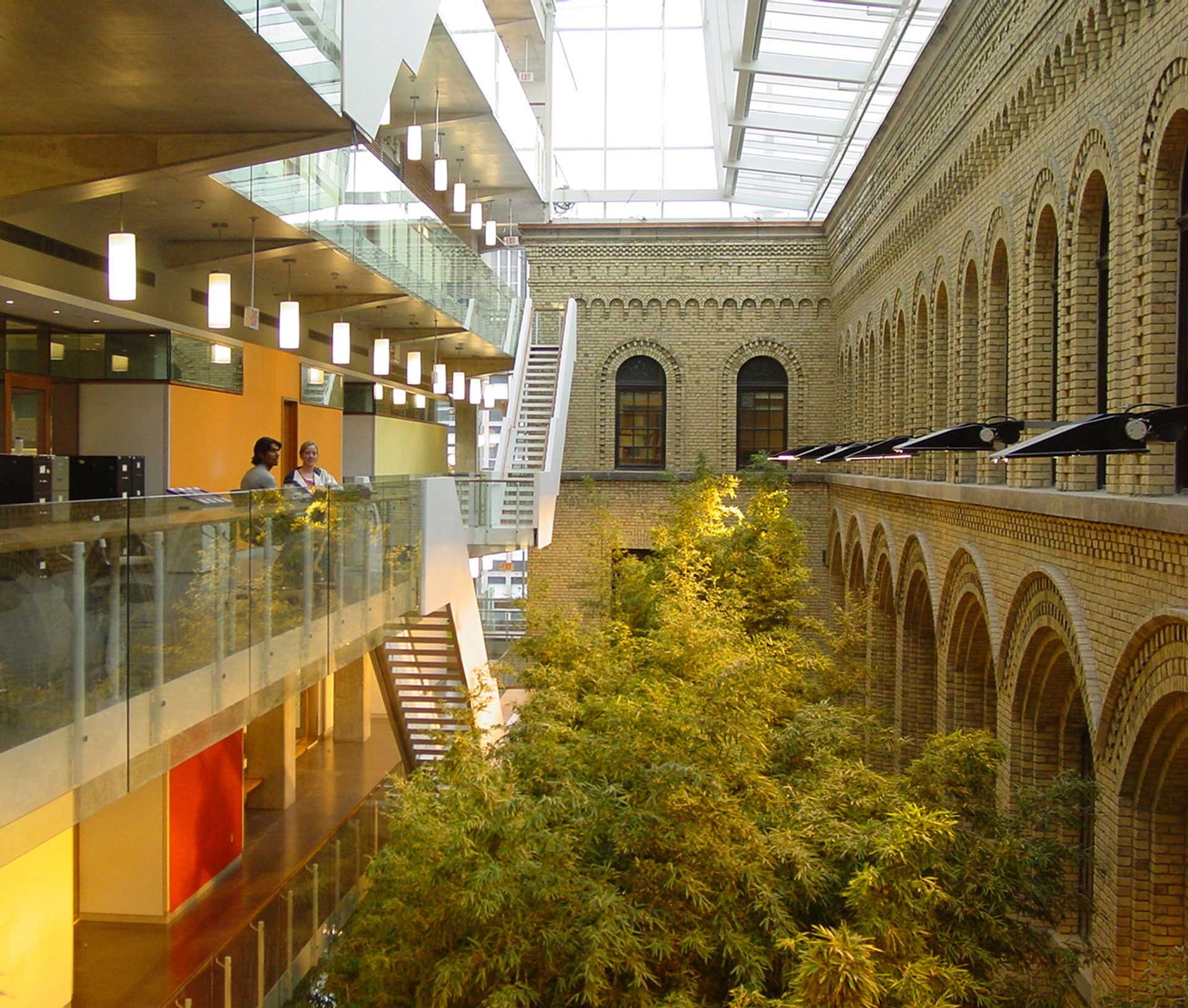 Project by Behnisch Architekturbuero, The Donnelly Centre for Cellular and Biomolecular Research. Interior atrium with  meeting areas and stairs, overlooking a planted courtyard, beside a brick facade with arched windows.