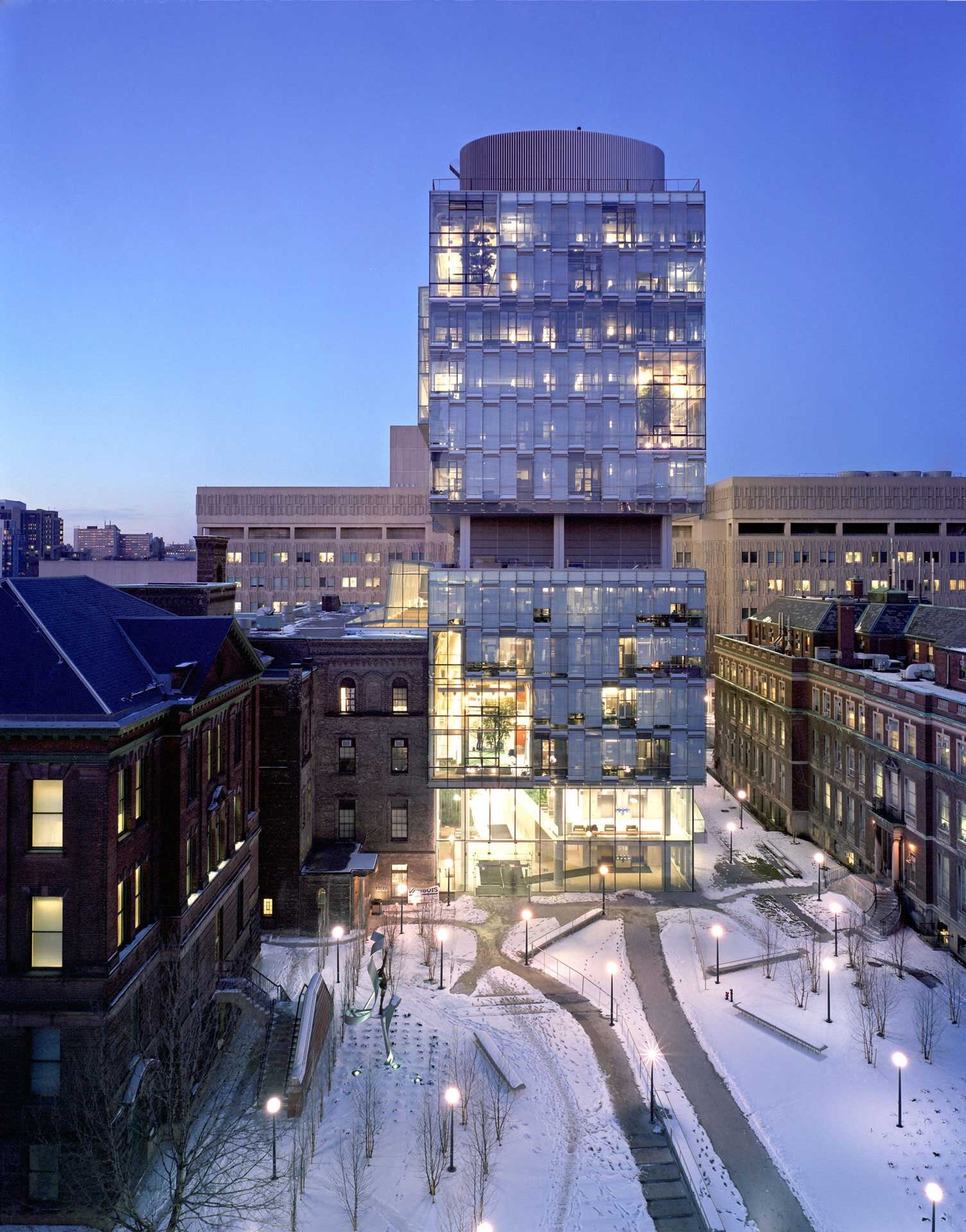 Project by Behnisch Architekturbuero, The Donnelly Centre for Cellular and Biomolecular Research. The illuminated glass high-rise building at dusk, surrounded by older buildings and a snowy courtyard with paths and streetlights.