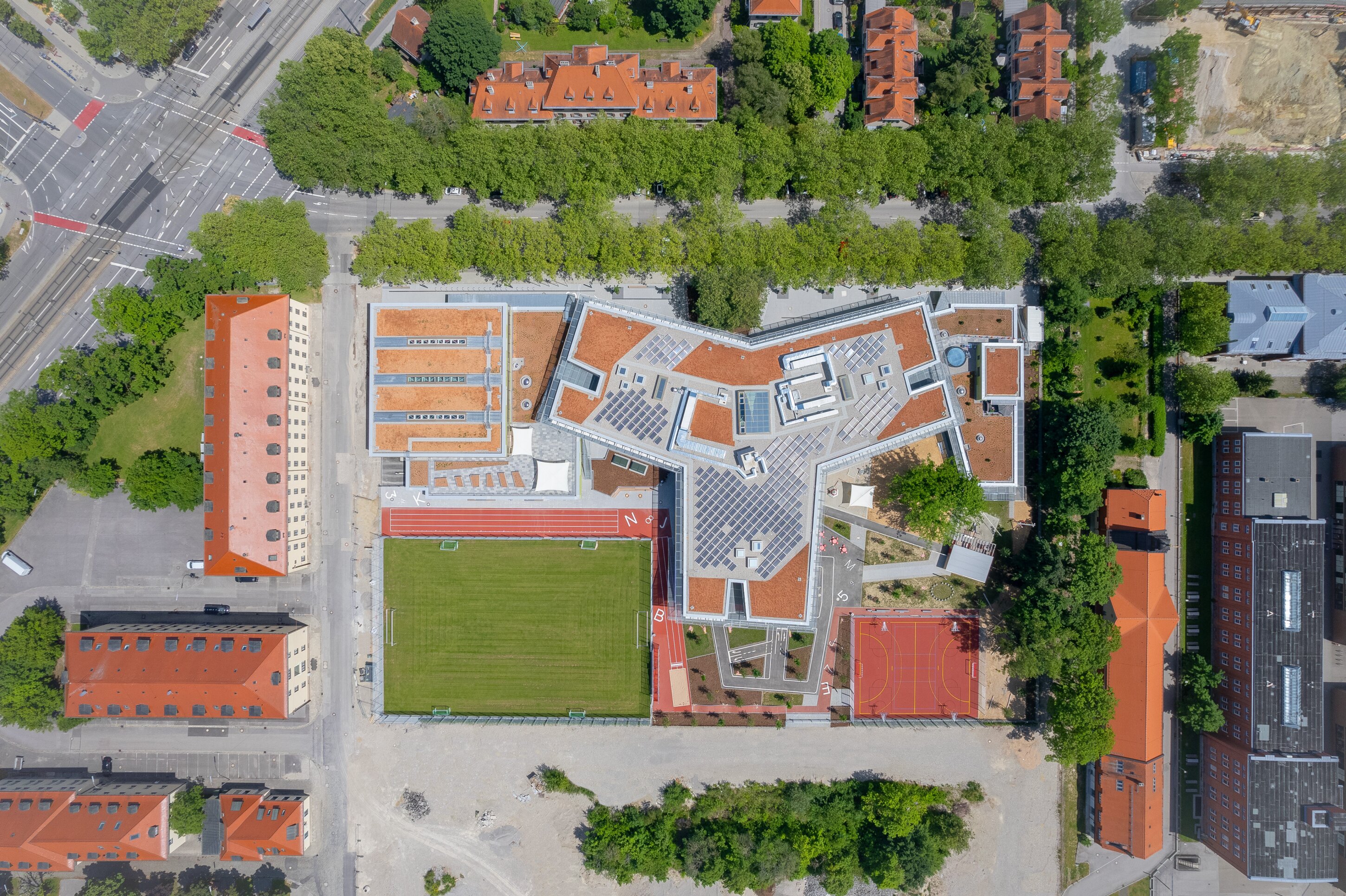 Project by Behnisch Architekturbuero, Primaryschool Infanteriestra&szlig;e. Aerial view of a school campus with buildings, green sports field, running track, courts, and surrounding streets and trees.
